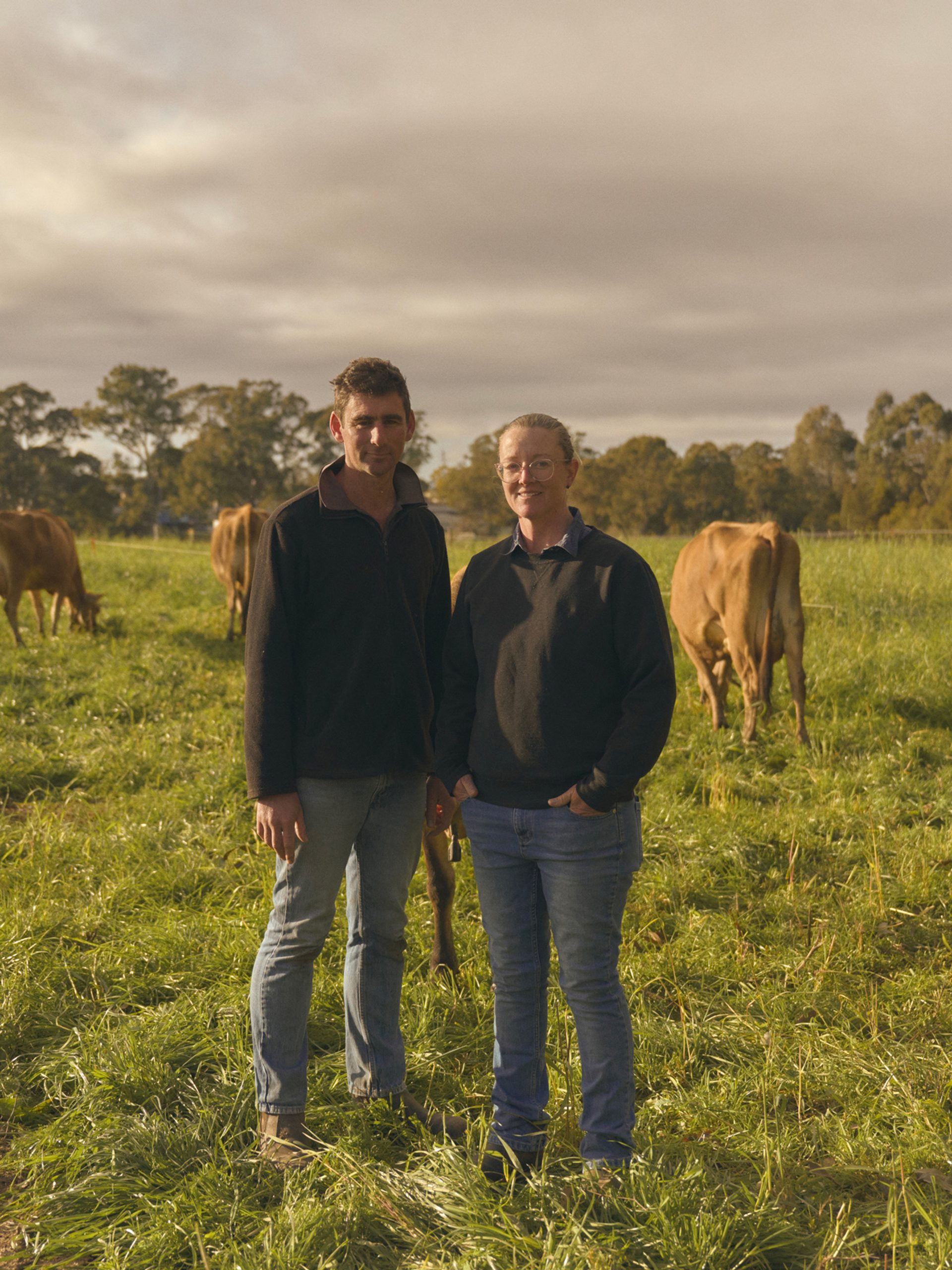 Two farmers stand side by side in a lush green pasture, surrounded by grazing cows. Both are dressed in casual outdoor clothing, with the man on the left wearing a dark jacket and jeans, and the woman on the right in a similar outfit. The cows, with their light brown coats, are calmly eating grass, and the scene is set under a partly cloudy sky, with trees visible in the background.