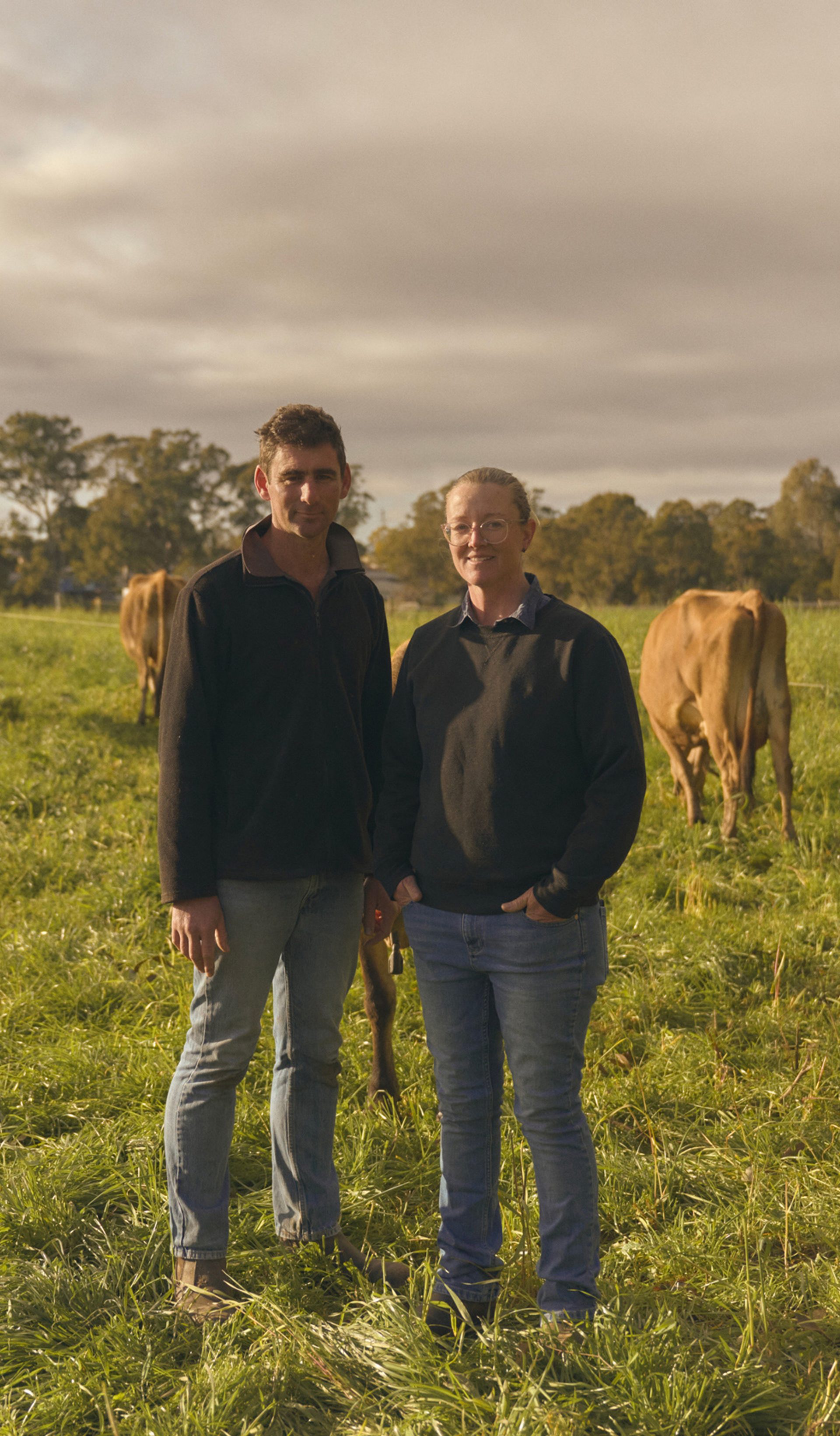 Two farmers stand side by side in a lush green pasture, surrounded by grazing cows. Both are dressed in casual outdoor clothing, with the man on the left wearing a dark jacket and jeans, and the woman on the right in a similar outfit. The cows, with their light brown coats, are calmly eating grass, and the scene is set under a partly cloudy sky, with trees visible in the background.