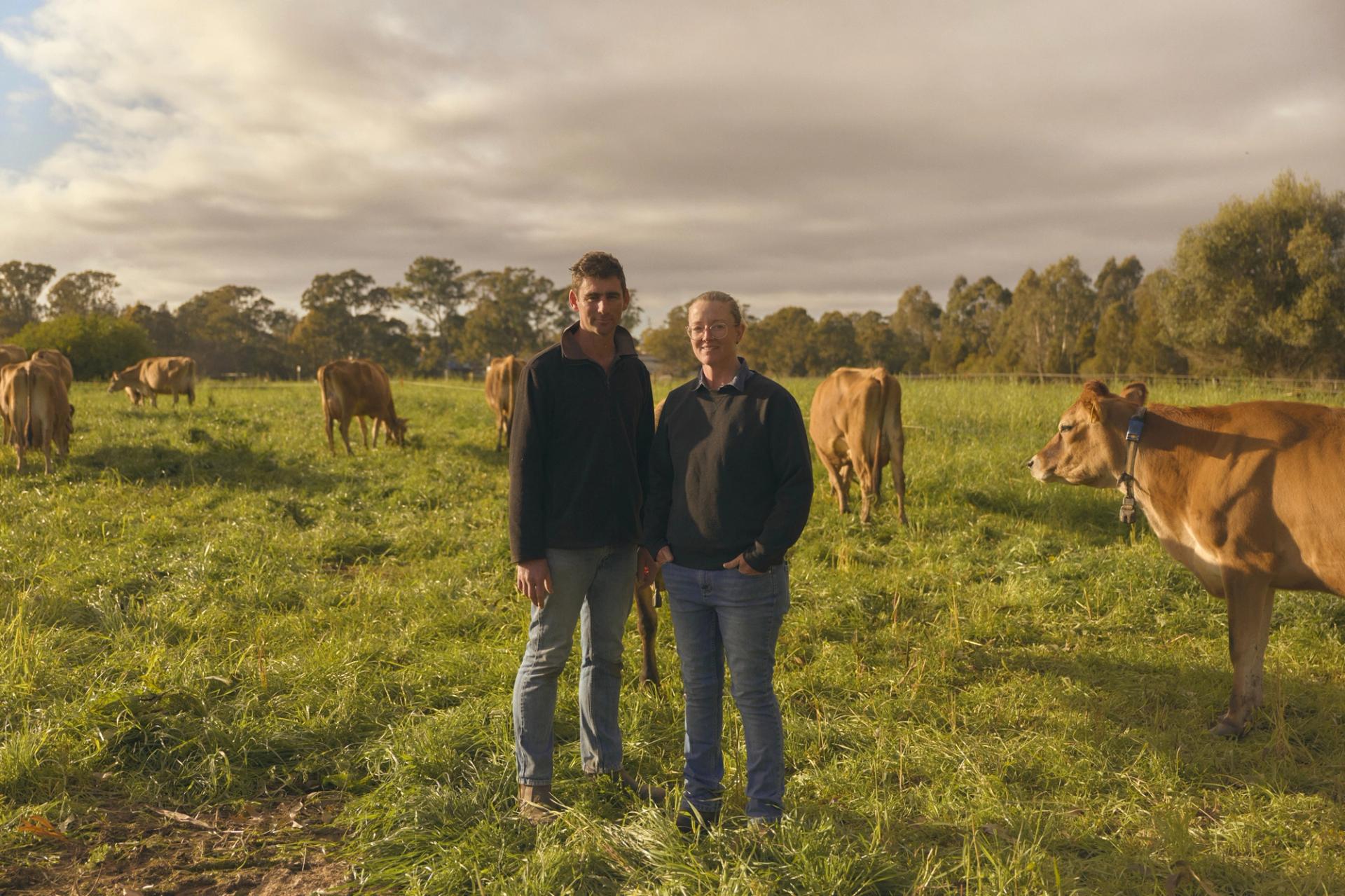 Two farmers stand side by side in a lush green pasture, surrounded by grazing cows. Both are dressed in casual outdoor clothing, with the man on the left wearing a dark jacket and jeans, and the woman on the right in a similar outfit. The cows, with their light brown coats, are calmly eating grass, and the scene is set under a partly cloudy sky, with trees visible in the background.
