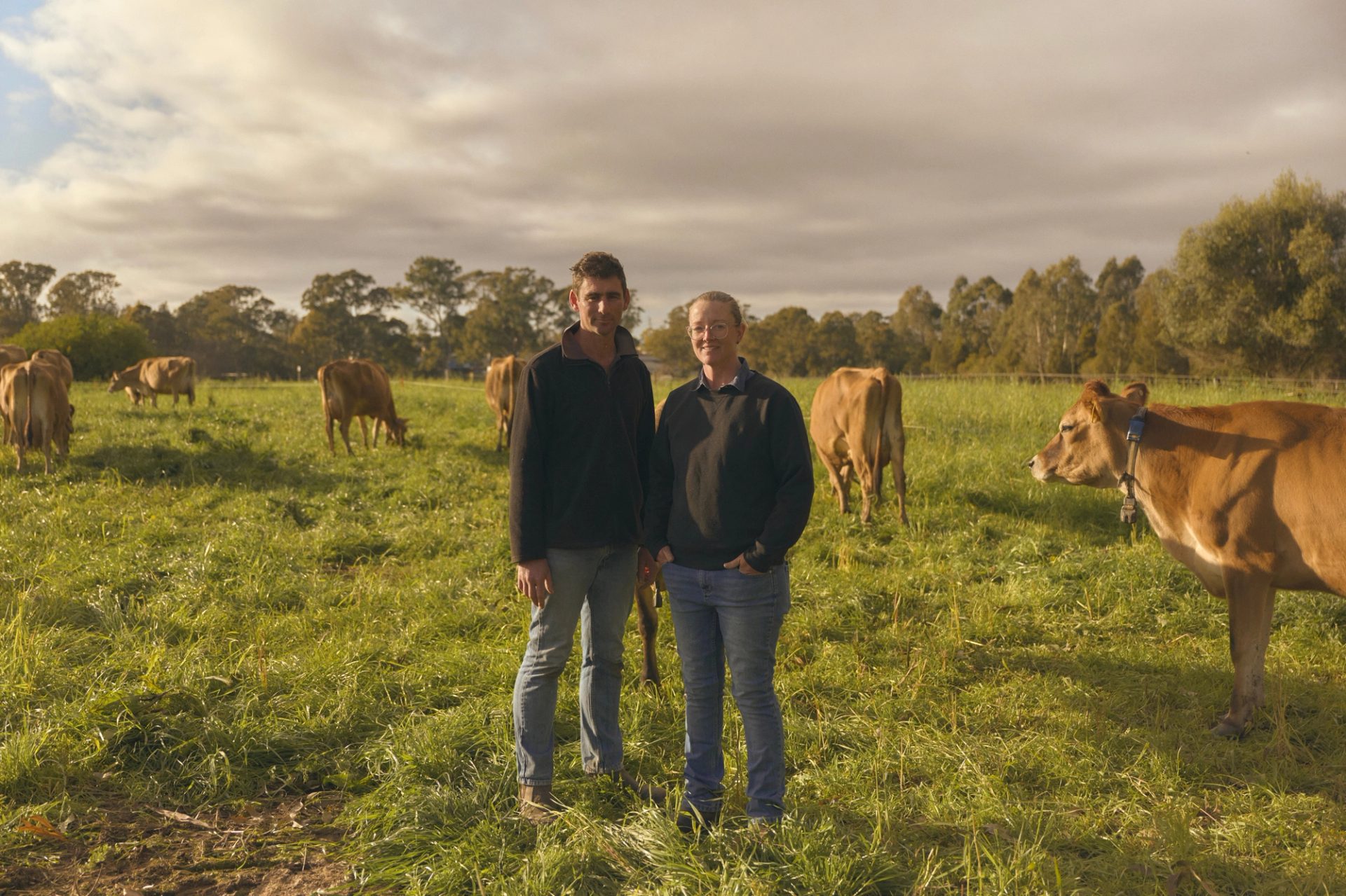 Two farmers stand side by side in a lush green pasture, surrounded by grazing cows. Both are dressed in casual outdoor clothing, with the man on the left wearing a dark jacket and jeans, and the woman on the right in a similar outfit. The cows, with their light brown coats, are calmly eating grass, and the scene is set under a partly cloudy sky, with trees visible in the background.