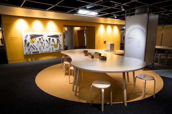 A large wooden table and stools in an exhibition space. On the far wall there is a board with a range of tools stuck to it.