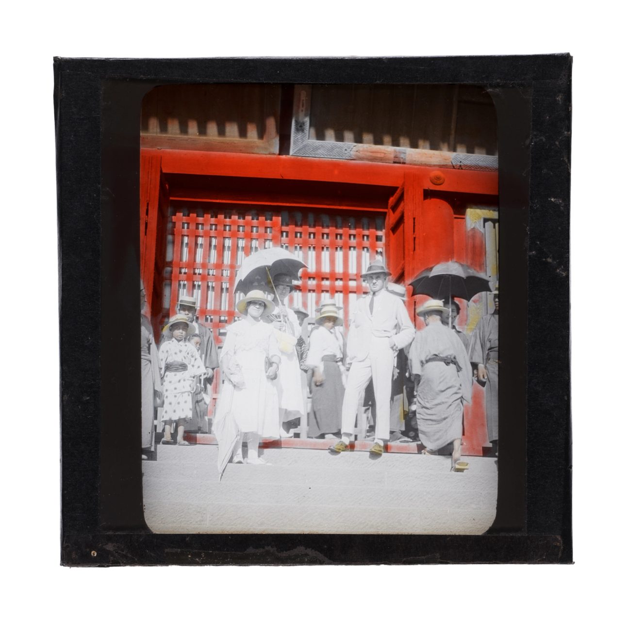 Silver gelatine glass plate negative in landscape orientation, depiciting in the foreground men, women and children standing at the top of stairs in front of a red gate. People wear a mixture of a white suit or dress and a kimono. Two people hold umbrellas.