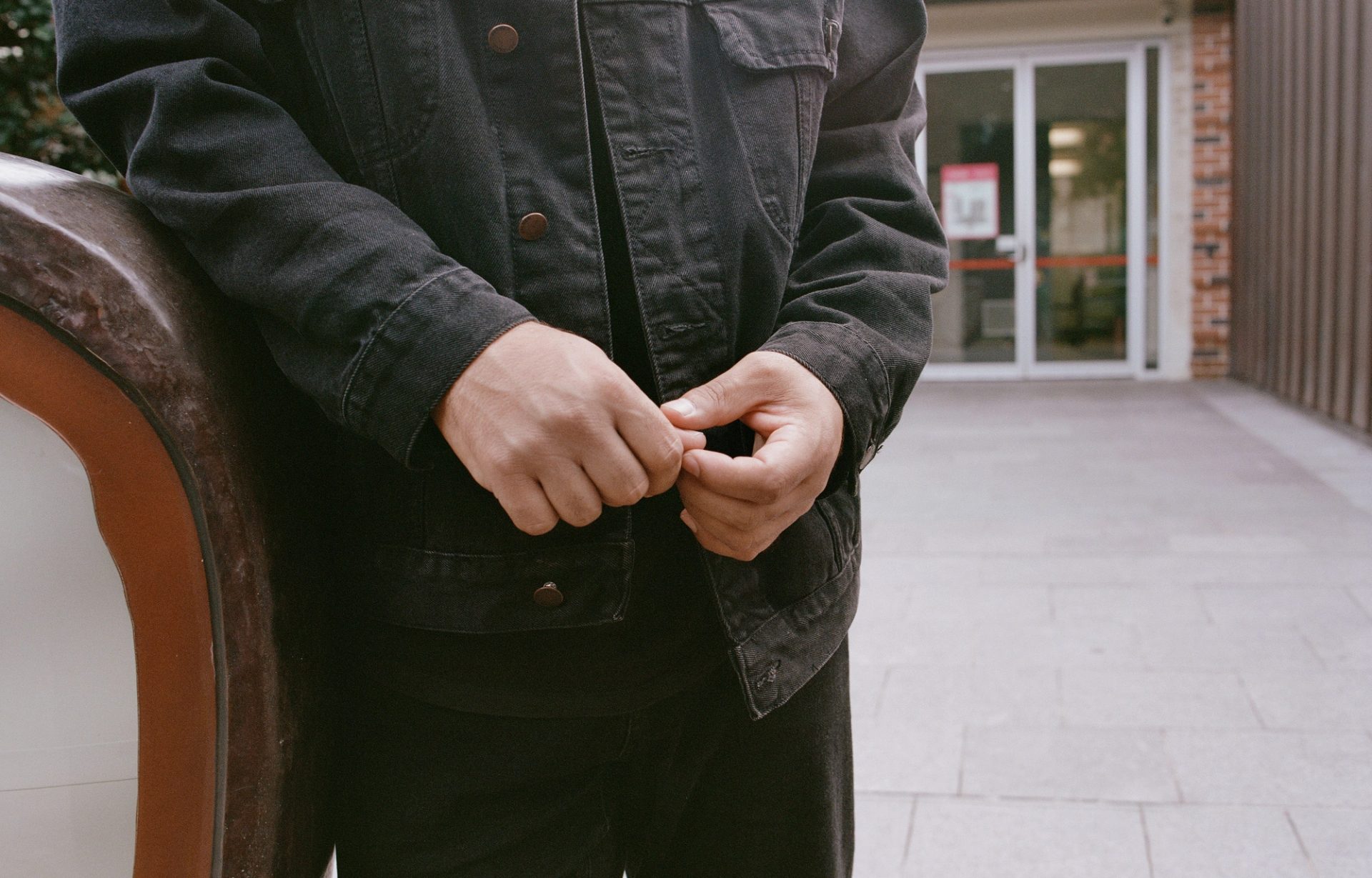 Person leans on an outdoor bench, hands together in front of them.