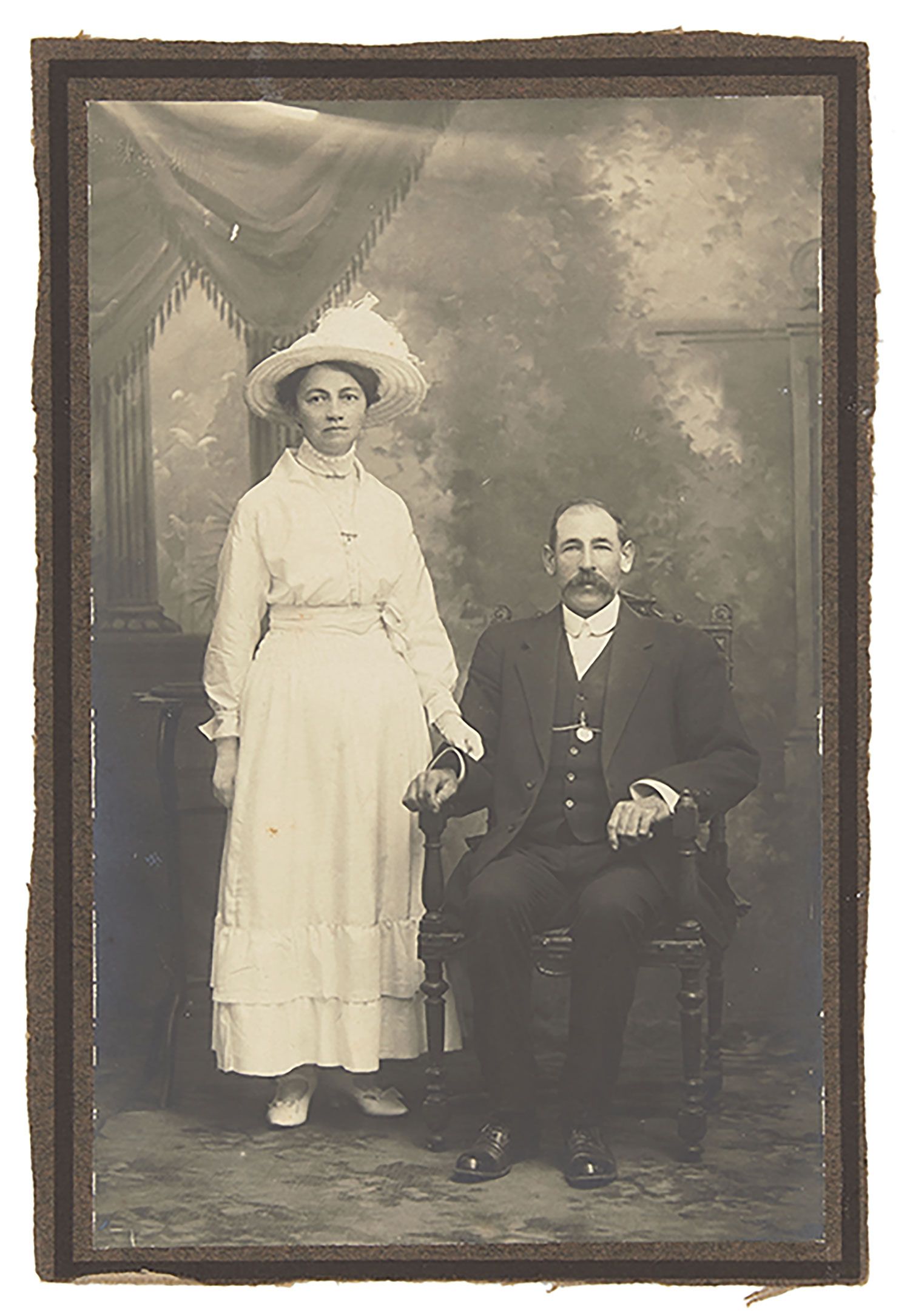 Mounted black and white wedding portrait photograph depicting a woman dressed in a full length white dress and white hat standing next to a seated man with a moustache and wearing a tuxedo.