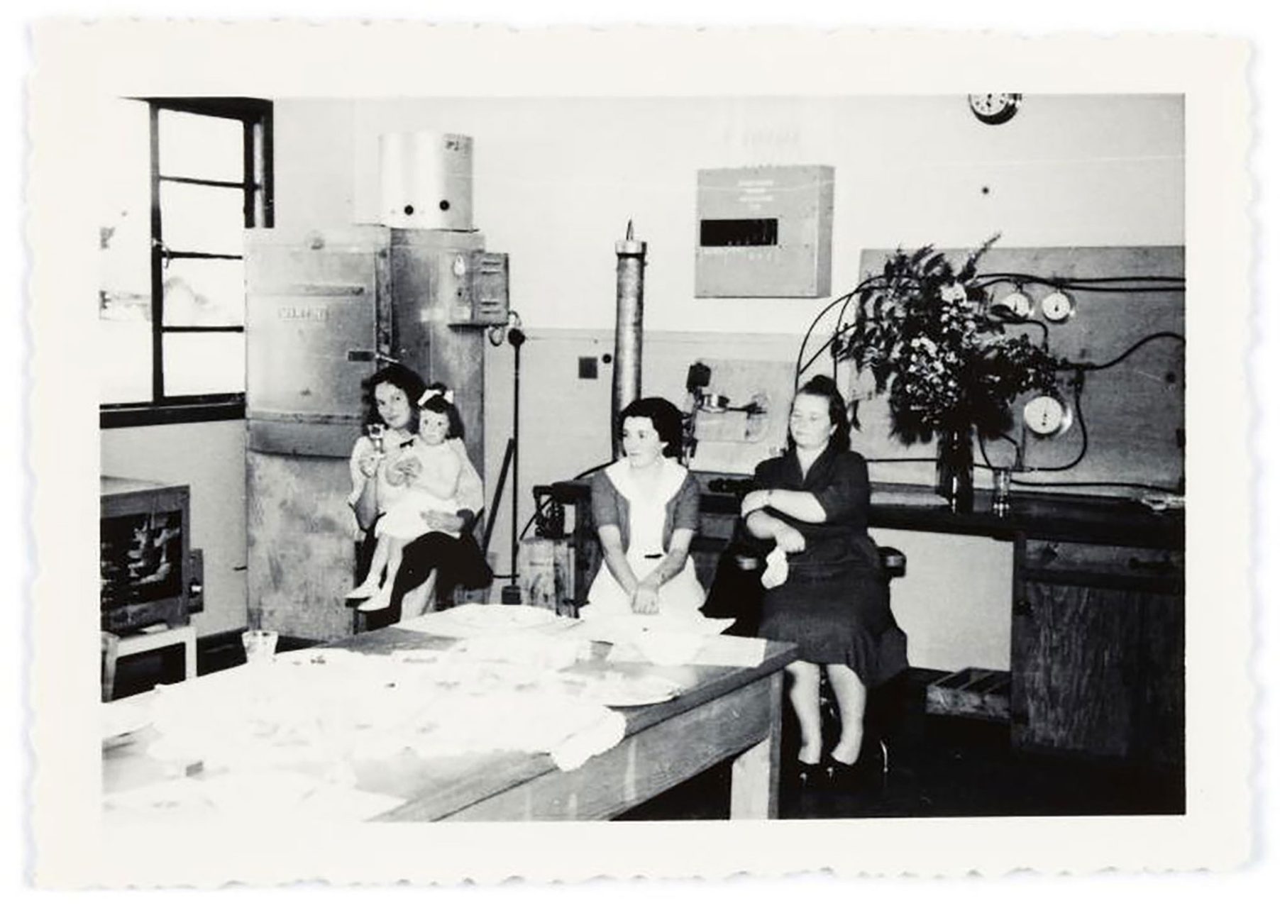 Black and white photograph of three women seated in a large kitchen. One of the women is holding a young girl.