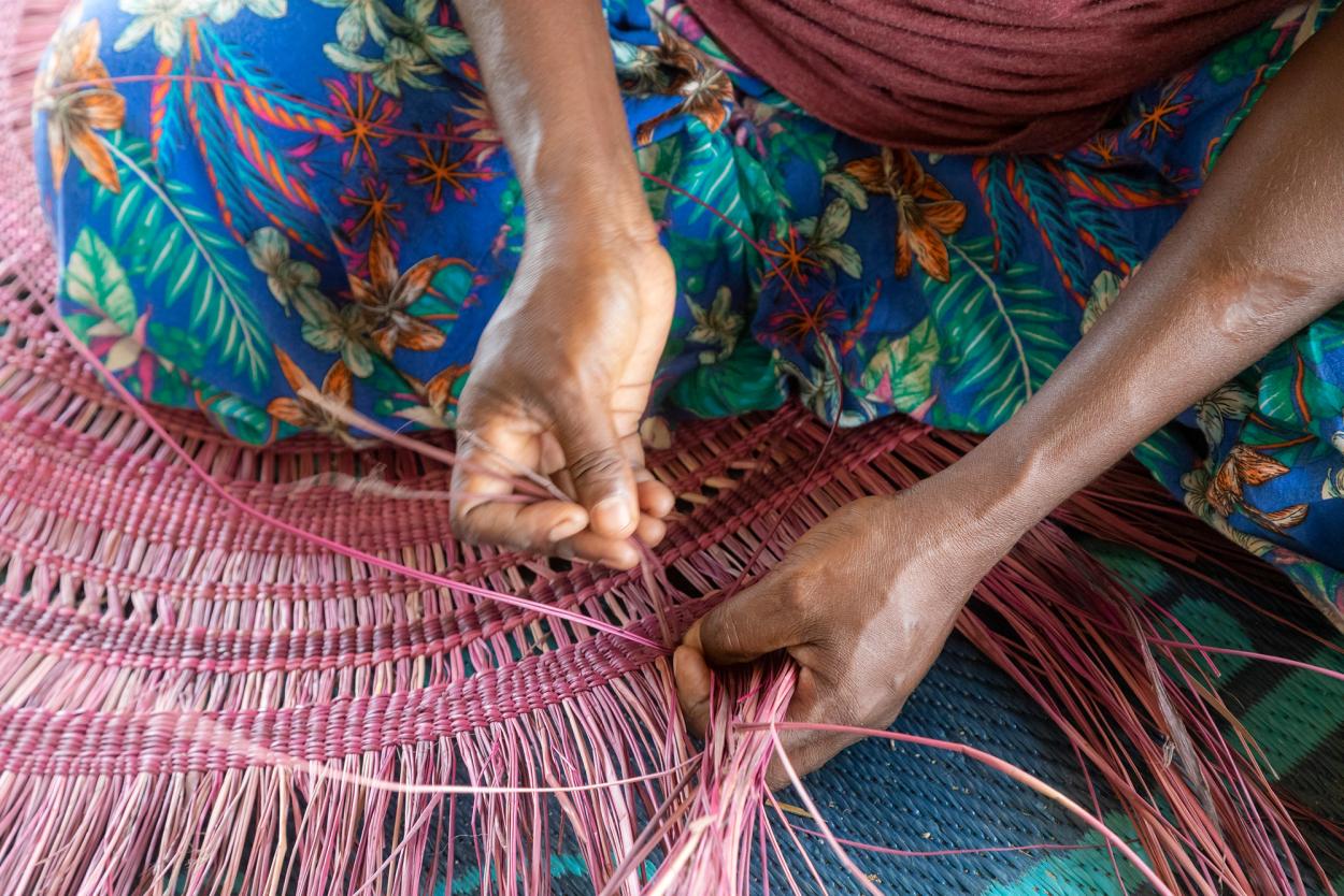 Brown hands of a woman stitching a pink traditional woven mat.
