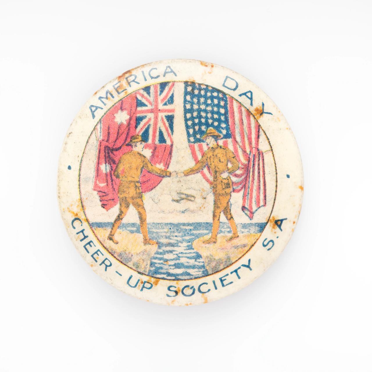 A badge made of plastic depicting an 'America Day - Cheer-up Society S.A' with an illustration of two soldiers shaking hands with the Australian and American flag behind each.