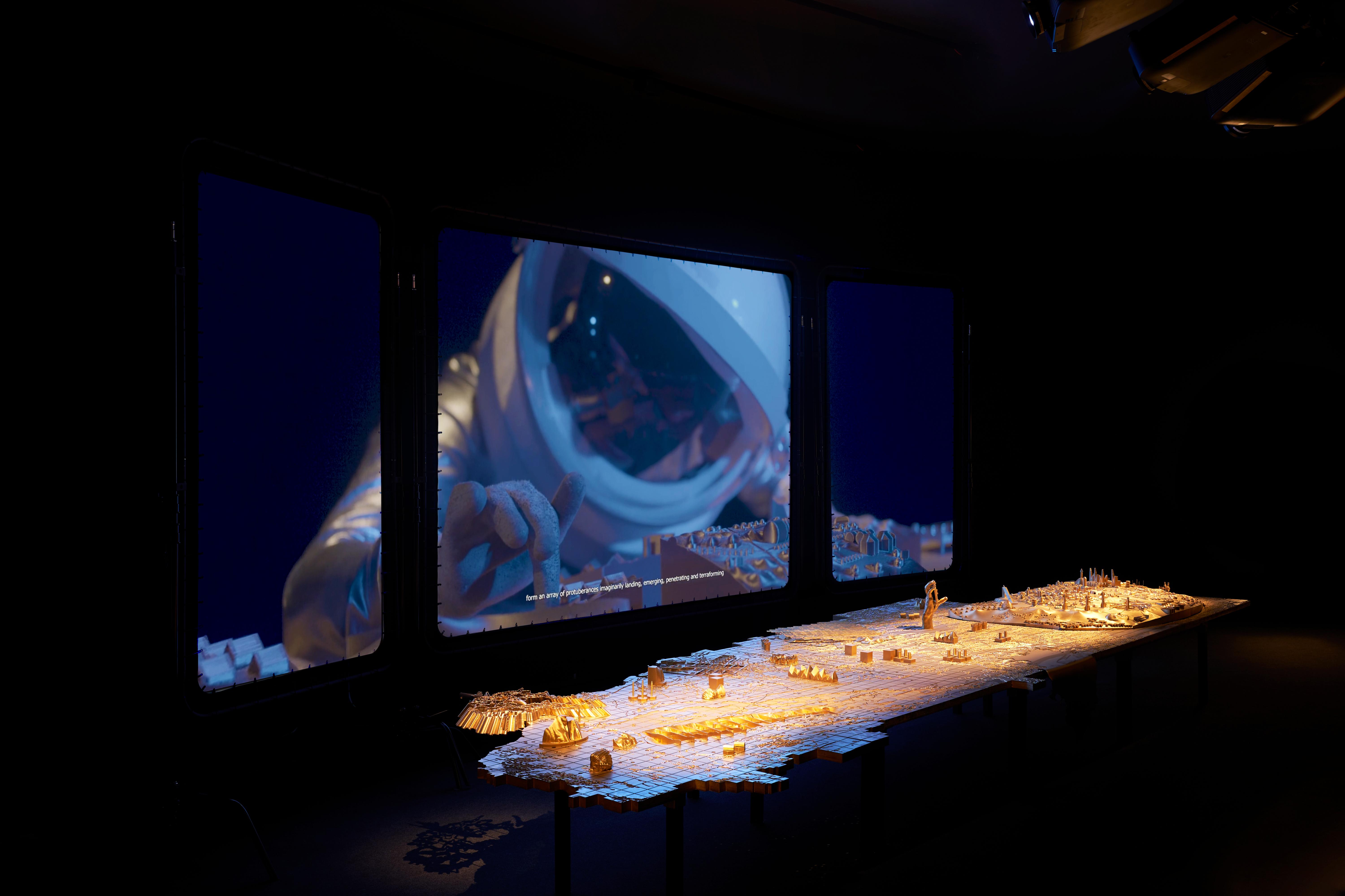 A long, golden display of abstract items sits in the foreground in a large dark space. Behind it is a large screen depicting an astronaut-like person.