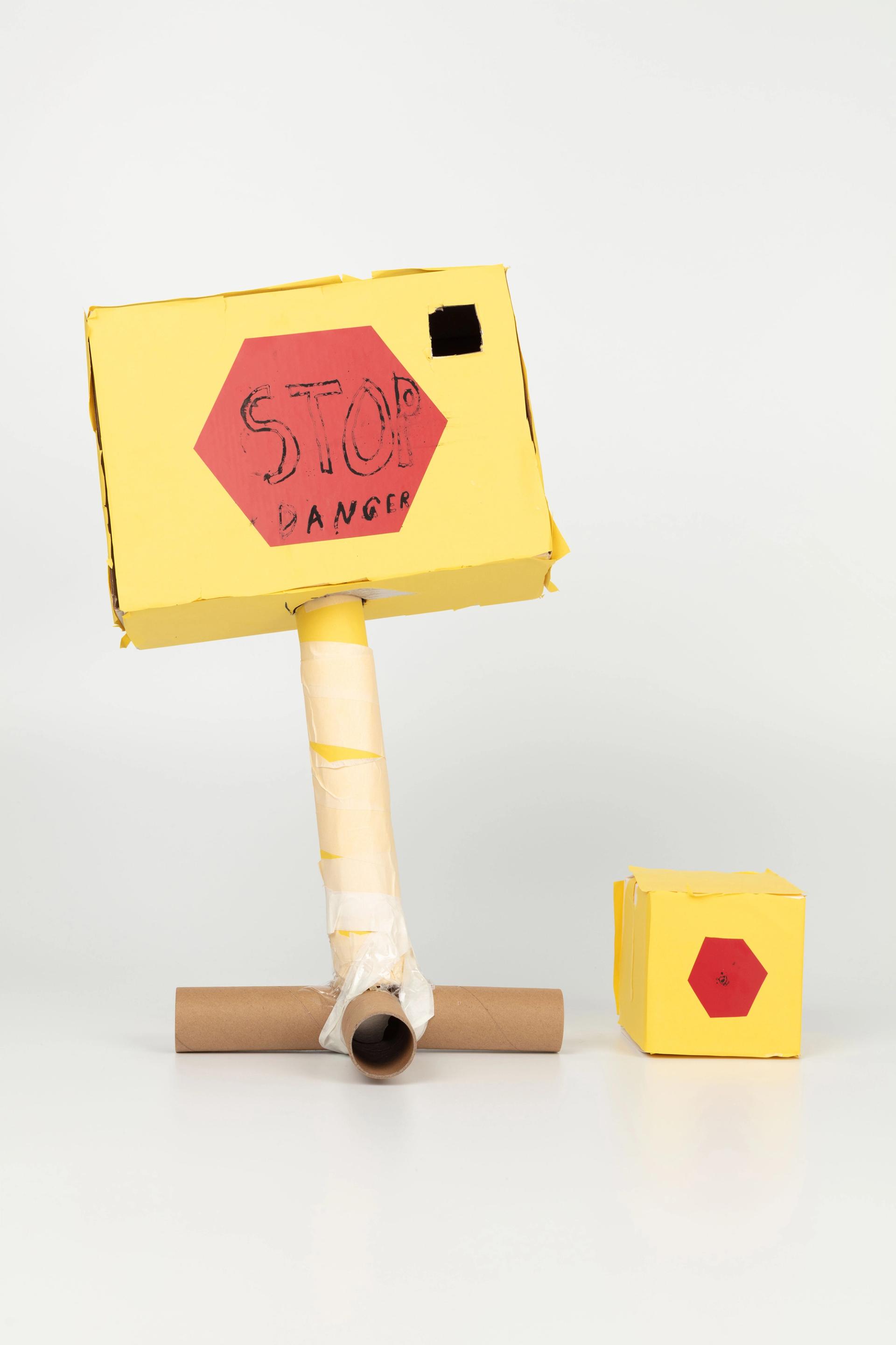 Image of a hand crafted mechanical device against a white background. A yellow box on top of cardboard rolls, with a red hexagon label with the words ‘STOP - DANGER’ stuck to the front. A smaller version of the cube sits beside it.