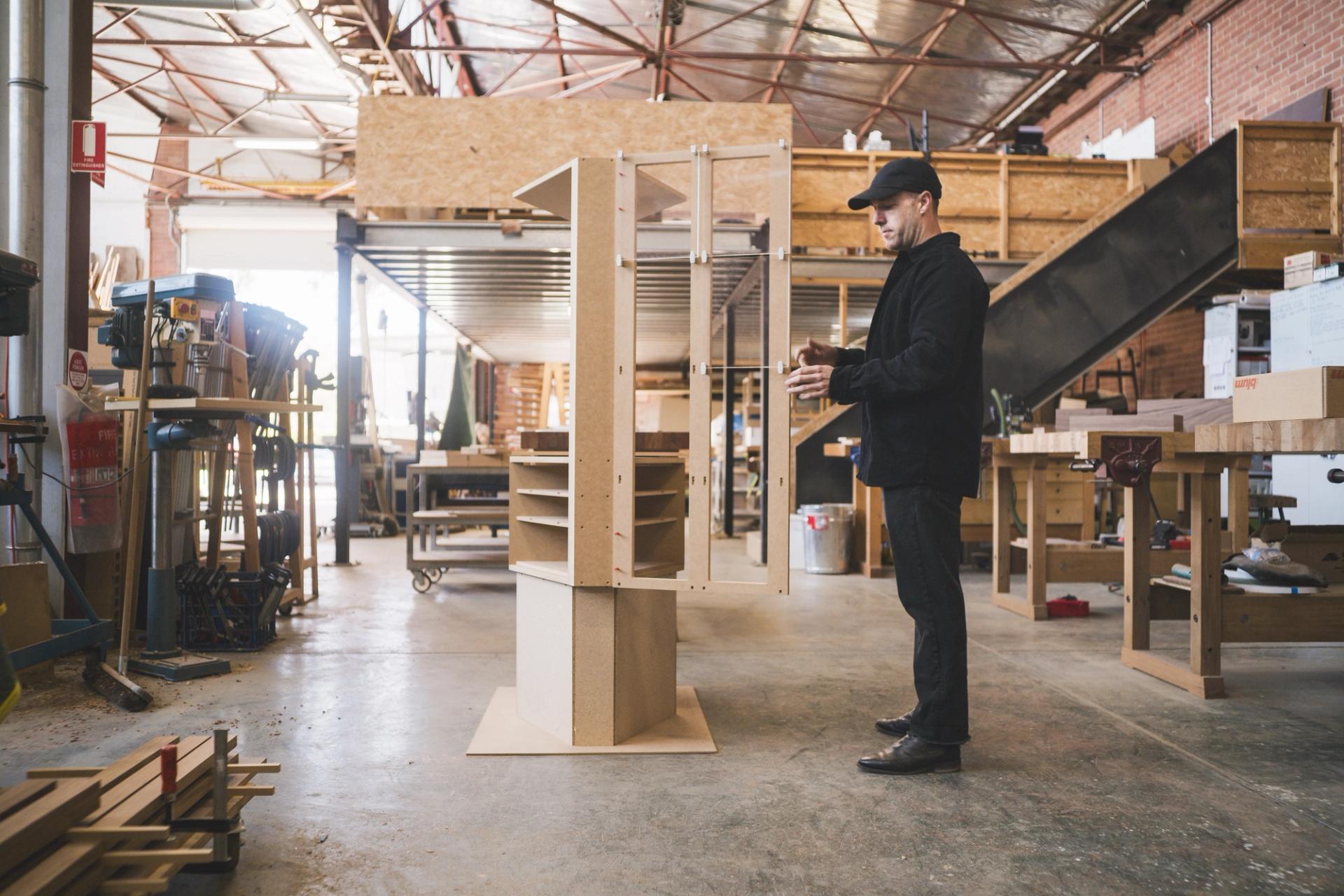 In a light-filled furniture workshop, a man interacts with a prototype of a large cabinet. He’s dressed in black.