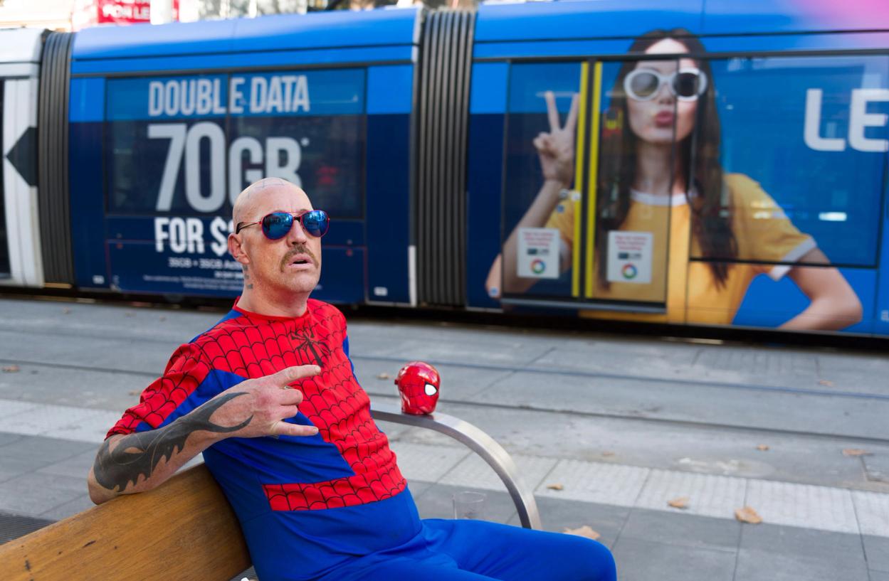 A figure with a handlebar moustache in a Spider Man costume sits on a bench. A tram is in the background.