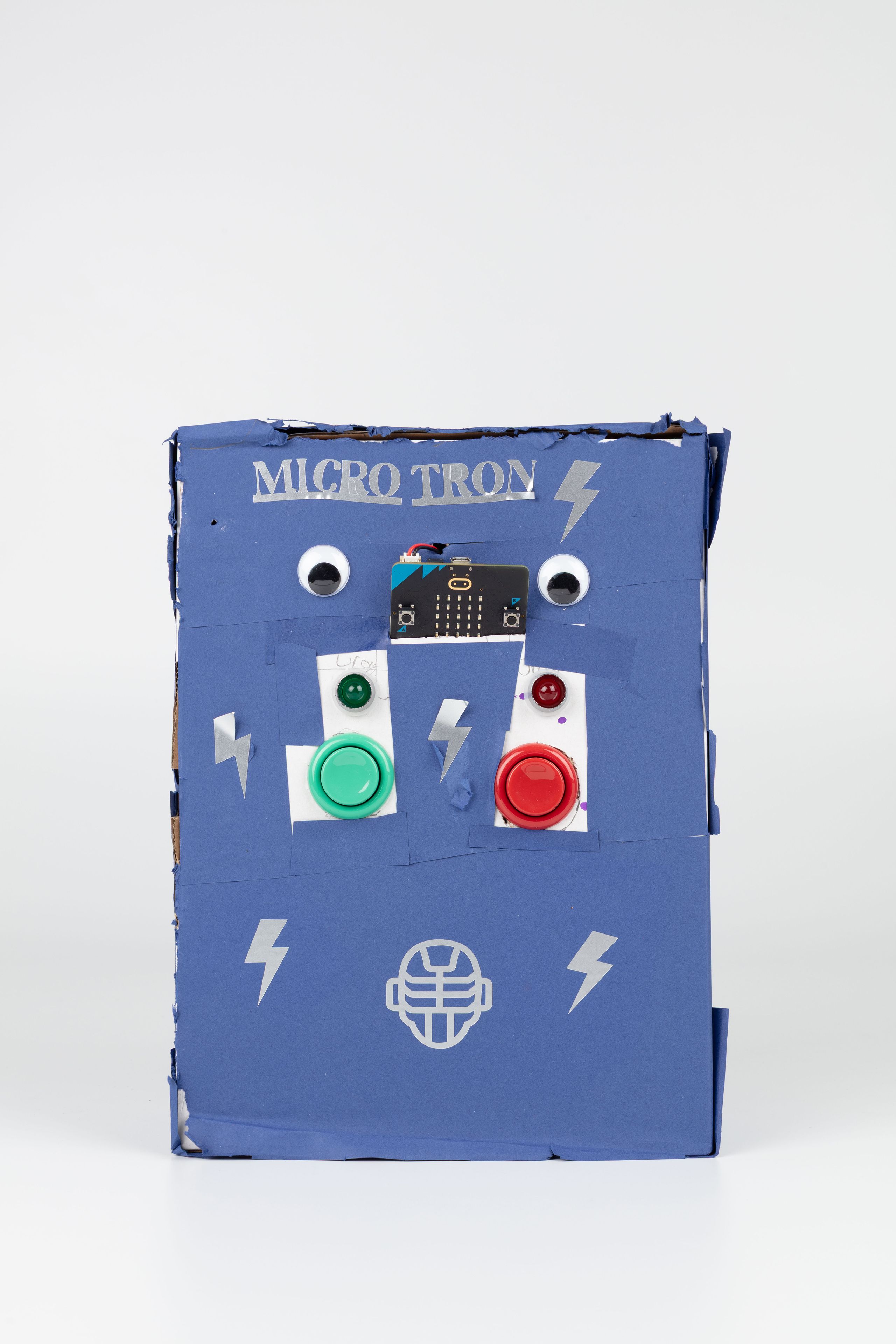 Image of a hand crafted mechanical device against a white background. A blue box decorated with lighting strike shapes and googly eyes, the words ‘MICRO TRON’ labelled on the top with a green and red button in the centre.