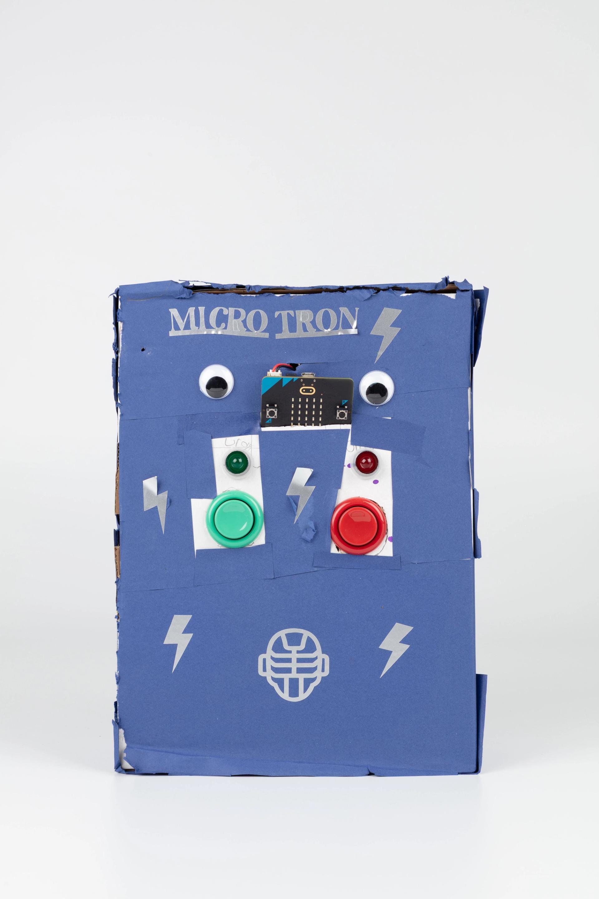 Image of a hand crafted mechanical device against a white background. A blue box decorated with lighting strike shapes and googly eyes, the words ‘MICRO TRON’ labelled on the top with a green and red button in the centre.