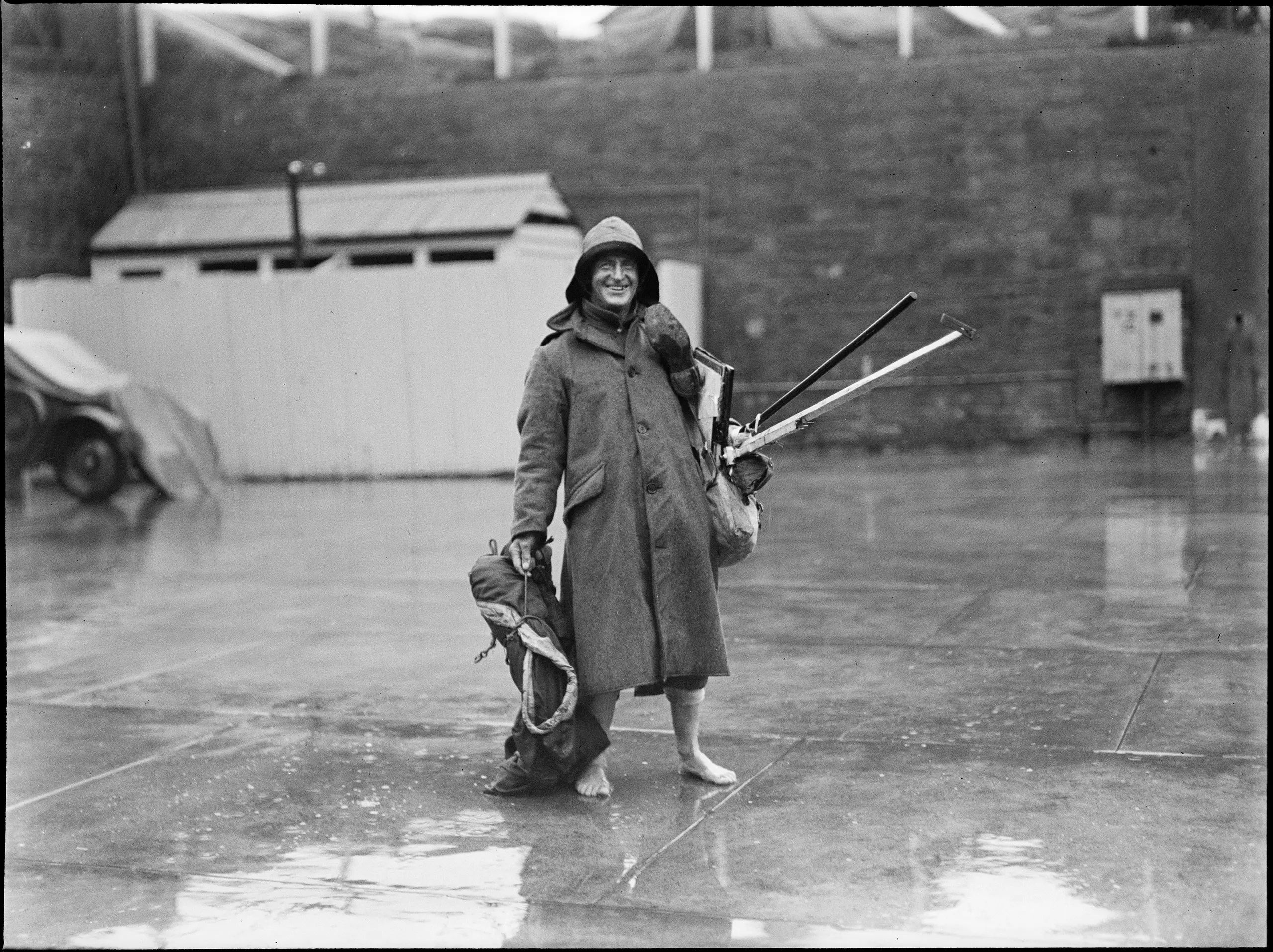 Glass negative, quarter plate, man in overcoat and souwester, Tom Lennon, Sydney, New South Wales, Australia, October 1933. Possibly at South Head Fort. The man is standing in a paved area where it has been raining. Behind him is a weatherboard and iron public toilet building and a high brick retaining wall. On the level above the wall, a tent may be seen pitched on the grass. The man is carrying equipment, possibly a tent and poles.