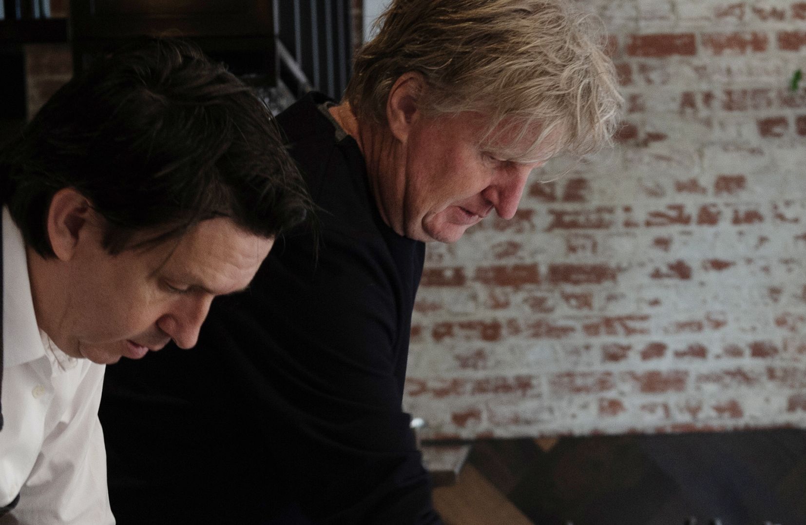 Two men looks down and over a table, in front of a brick wall.