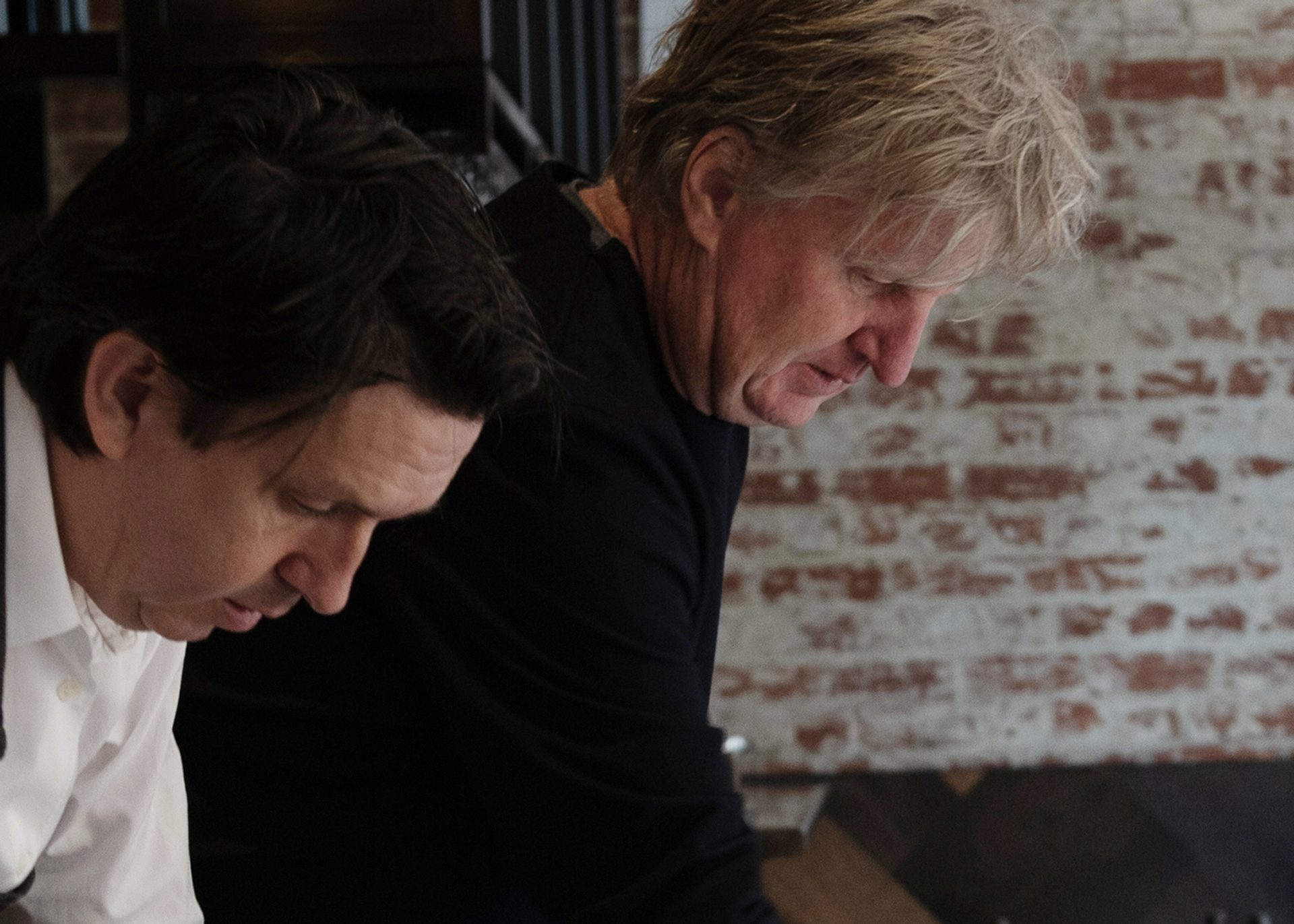 Two men looks down and over a table, in front of a brick wall.