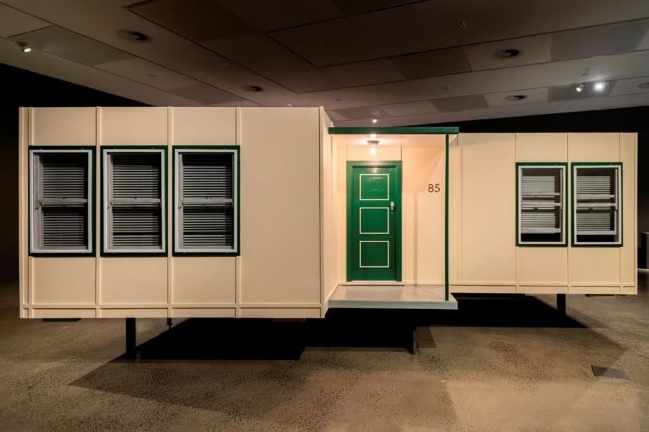 The sculpture is a 75-percent scale representation of front facade of pastel yellow fibro house with the number '85', though without the silhouette of the roofs. The panels contain no asbestos. Rectangular volumes that make up the sculpture are off-set, from each direction creating the effect of an L-shaped fibro house with a single-posted porch and a projecting front room.