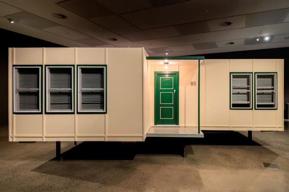 The sculpture is a 75-percent scale representation of front facade of pastel yellow fibro house with the number '85', though without the silhouette of the roofs. The panels contain no asbestos. Rectangular volumes that make up the sculpture are off-set, from each direction creating the effect of an L-shaped fibro house with a single-posted porch and a projecting front room.