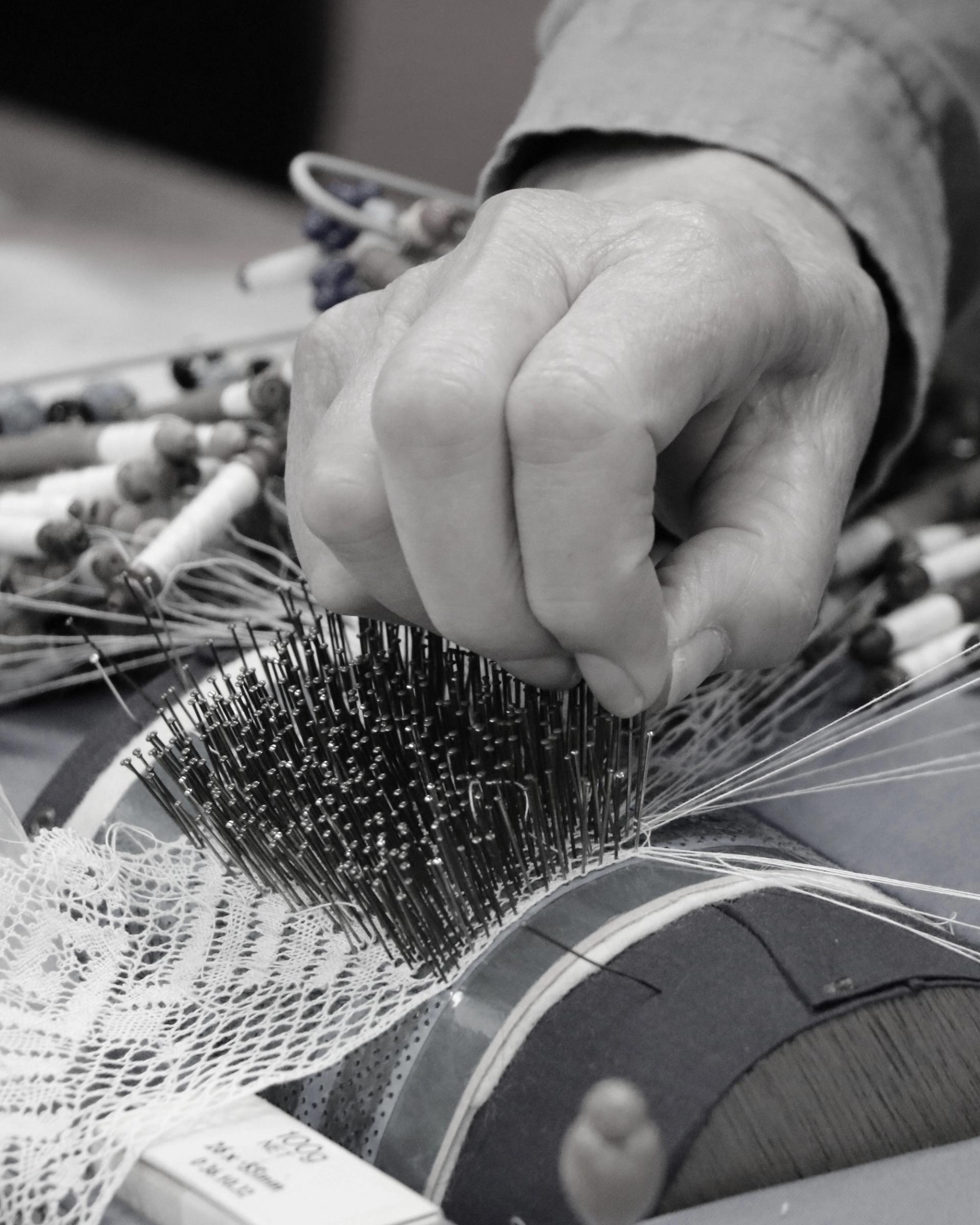 Black and white image of the lacemaking process, a hand pinching multiple pins onto a bobbin lacemaking machine to create a roll of lace.