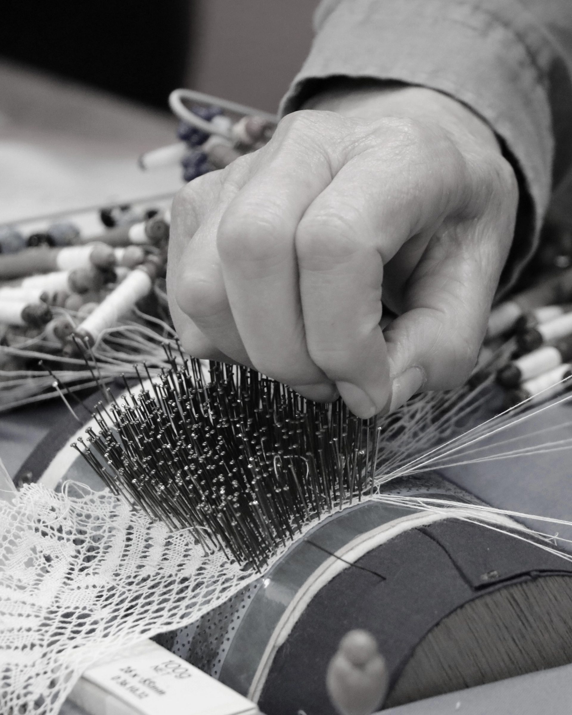 Black and white image of the lacemaking process, a hand pinching multiple pins onto a bobbin lacemaking machine to create a roll of lace.