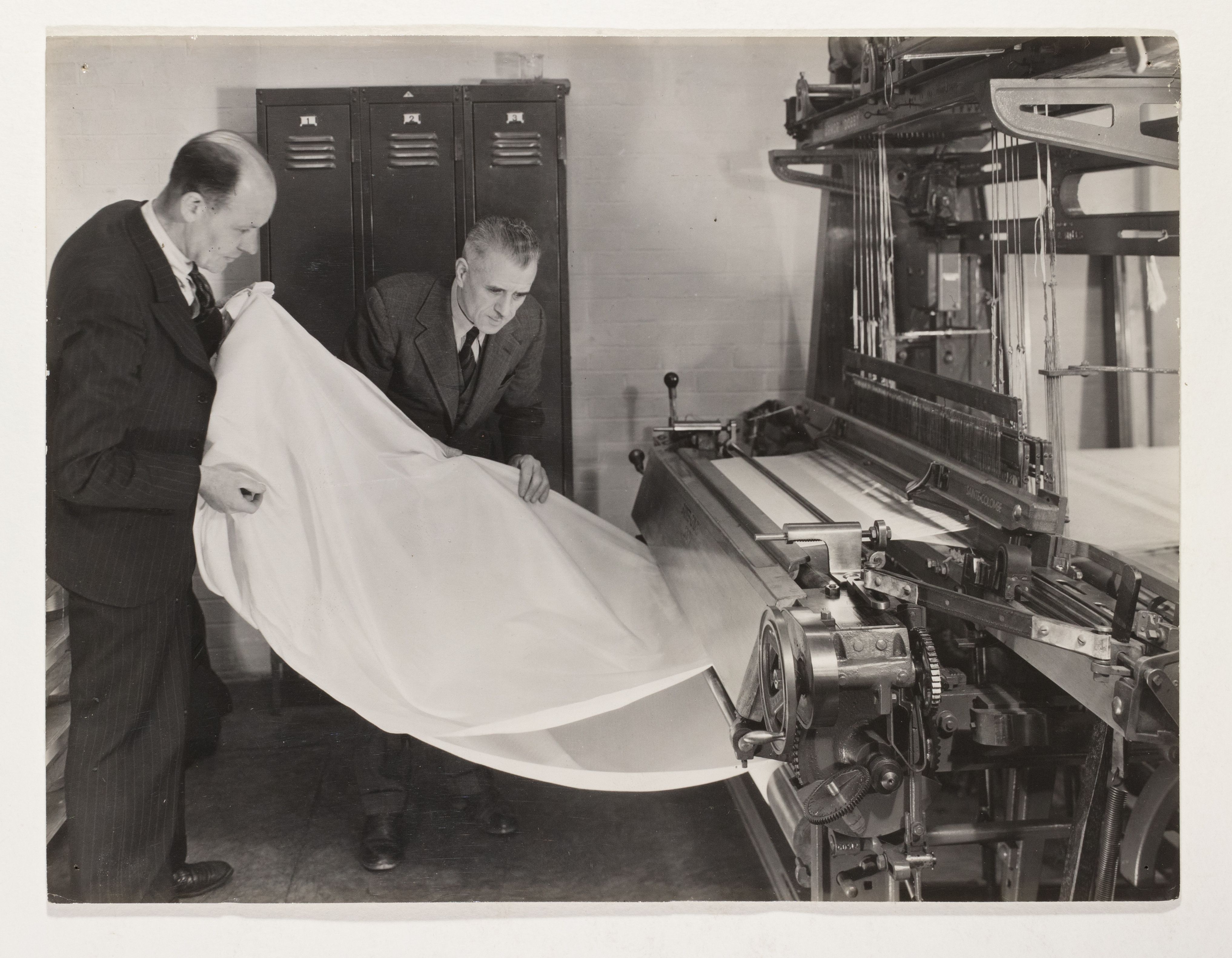 Black and white photograph depicting two men in suits examining a white fabric ('Terylene' synthetic material) being woven on an experimental loom.