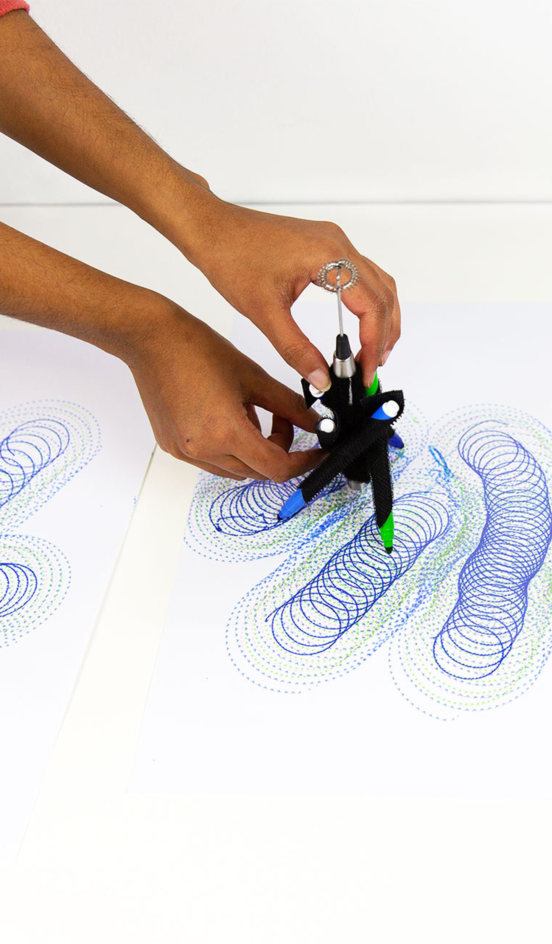 A series of spirograph drawings in blue and purple with hands controlling a DrawBot device.