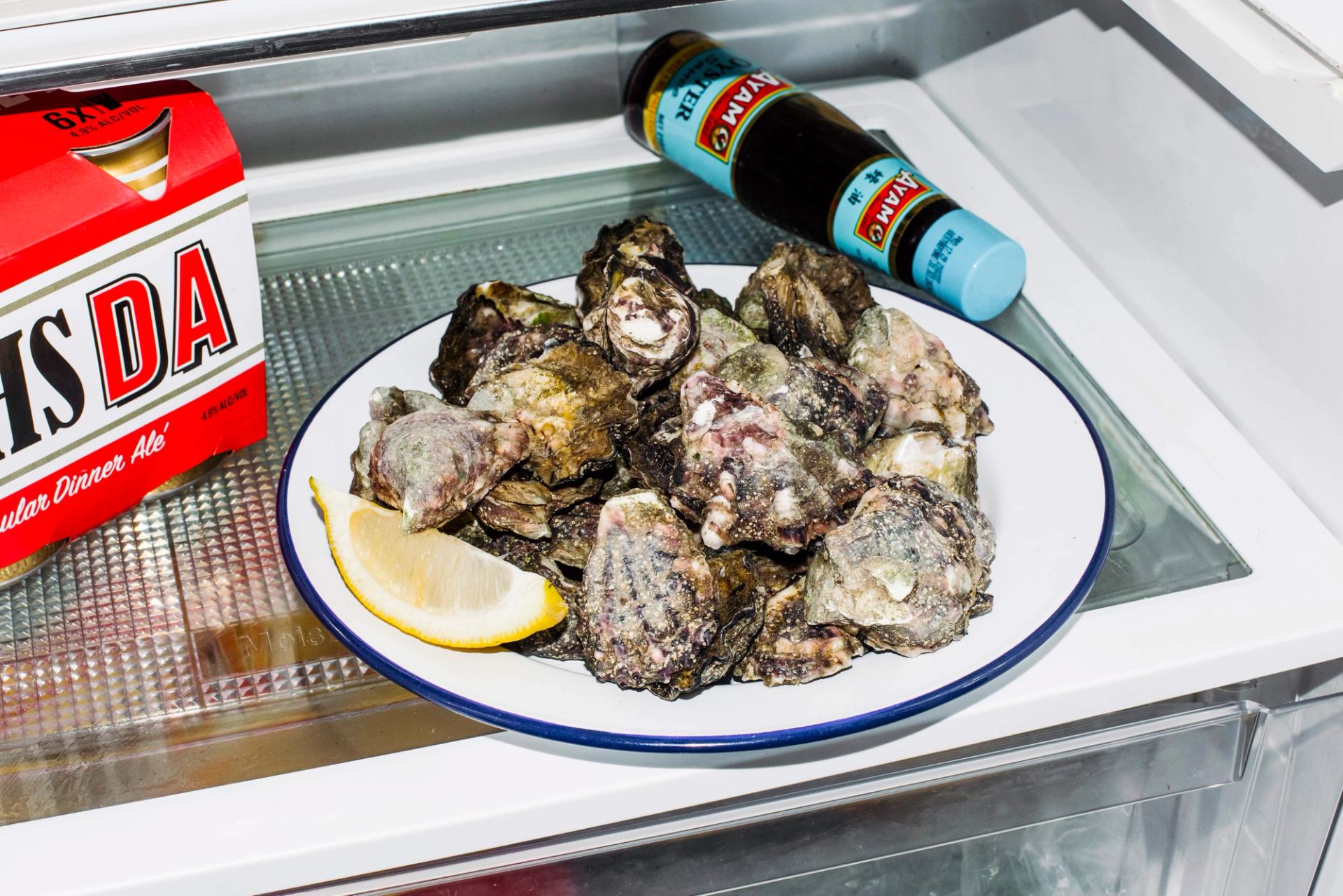 A plate of unchecked oysters with a slice of lemon on a white plate in a fridge.
