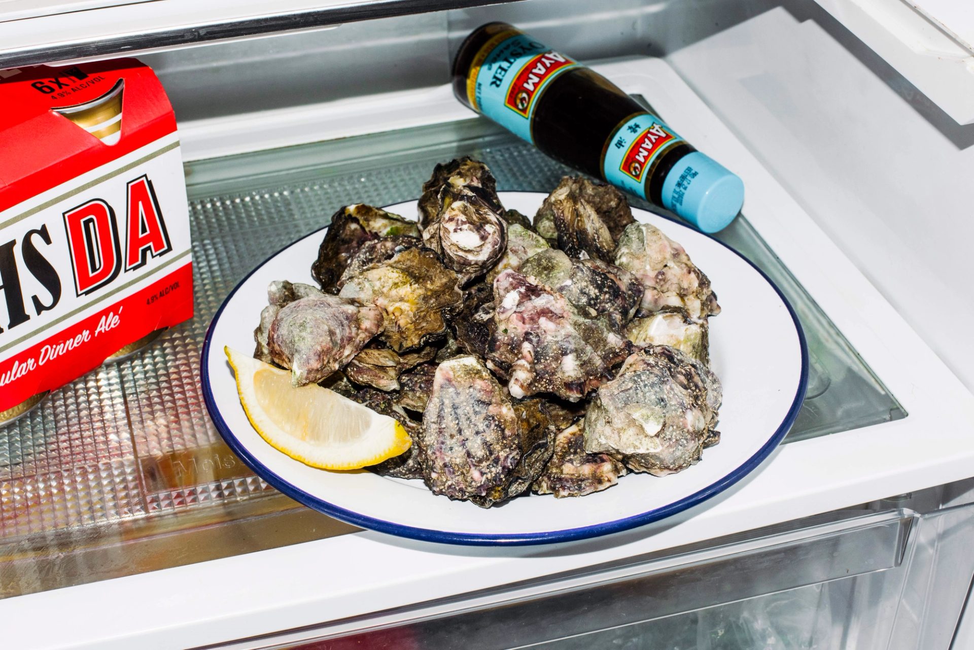 A plate of unchecked oysters with a slice of lemon on a white plate in a fridge.