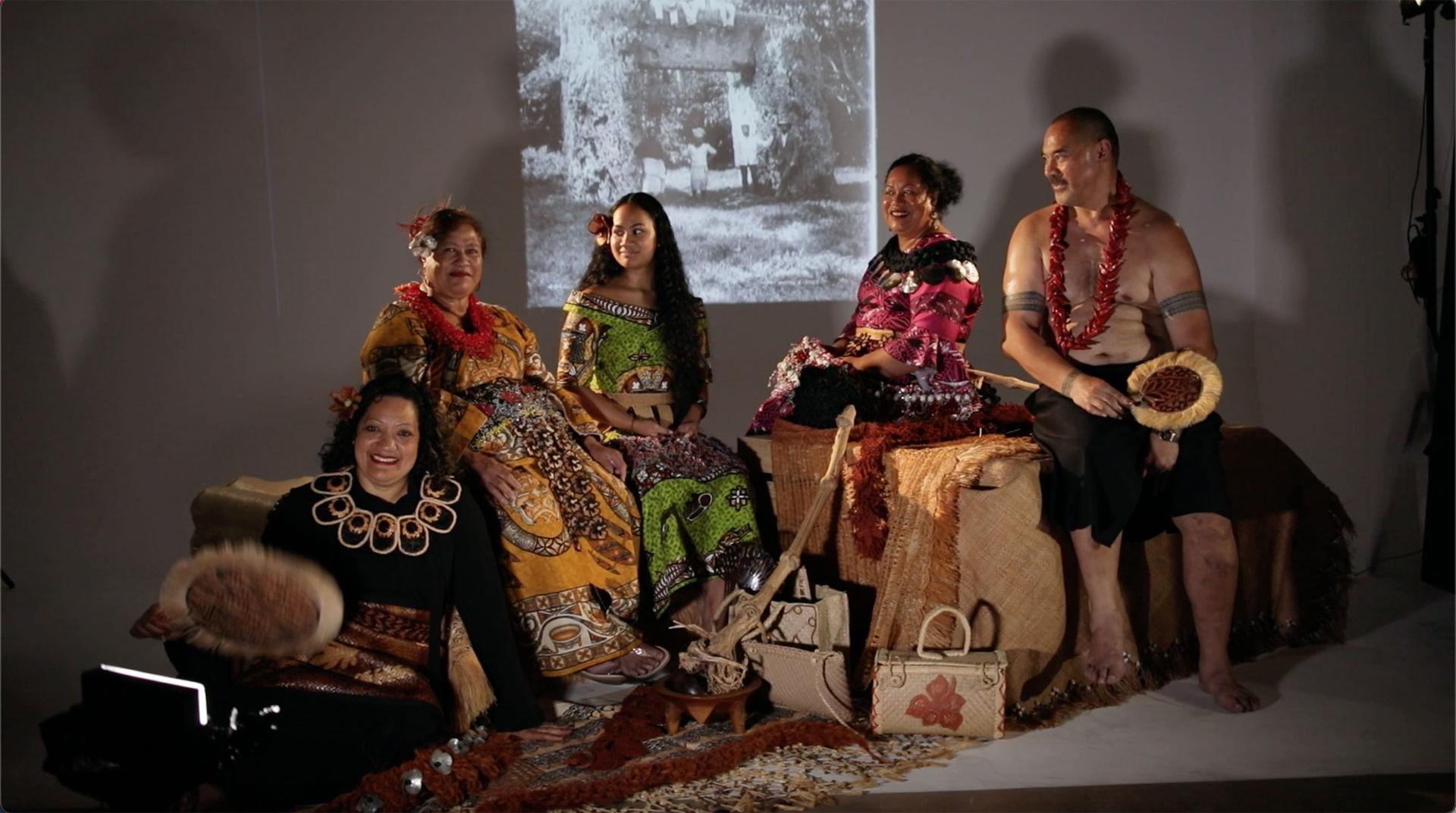 Four women and a man sitting in a semi-circle formation. One woman is sitting on the ground and the rest of the group are sitting on large boxes. They women are wearing traditional Tongan dress. The man is wearing traditional Samoan dress. They are holding a variety of objects.