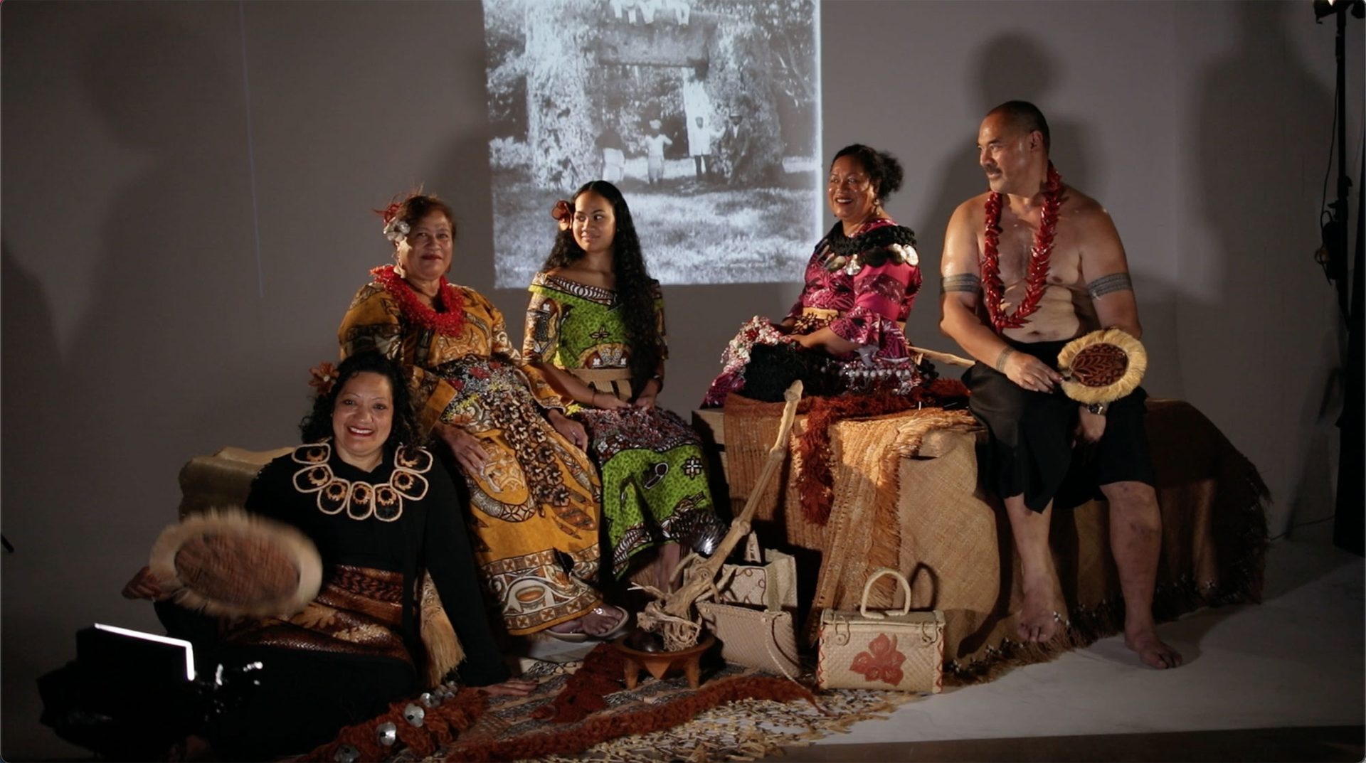 Four women and a man sitting in a semi-circle formation. One woman is sitting on the ground and the rest of the group are sitting on large boxes. They women are wearing traditional Tongan dress. The man is wearing traditional Samoan dress. They are holding a variety of objects.