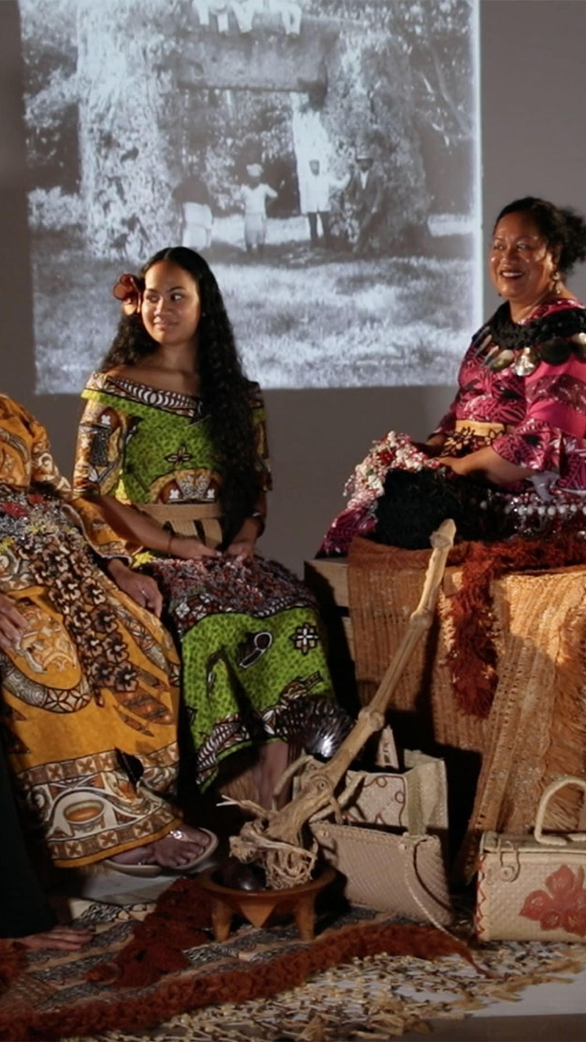 Four women and a man sitting in a semi-circle formation. One woman is sitting on the ground and the rest of the group are sitting on large boxes. They women are wearing traditional Tongan dress. The man is wearing traditional Samoan dress. They are holding a variety of objects.
