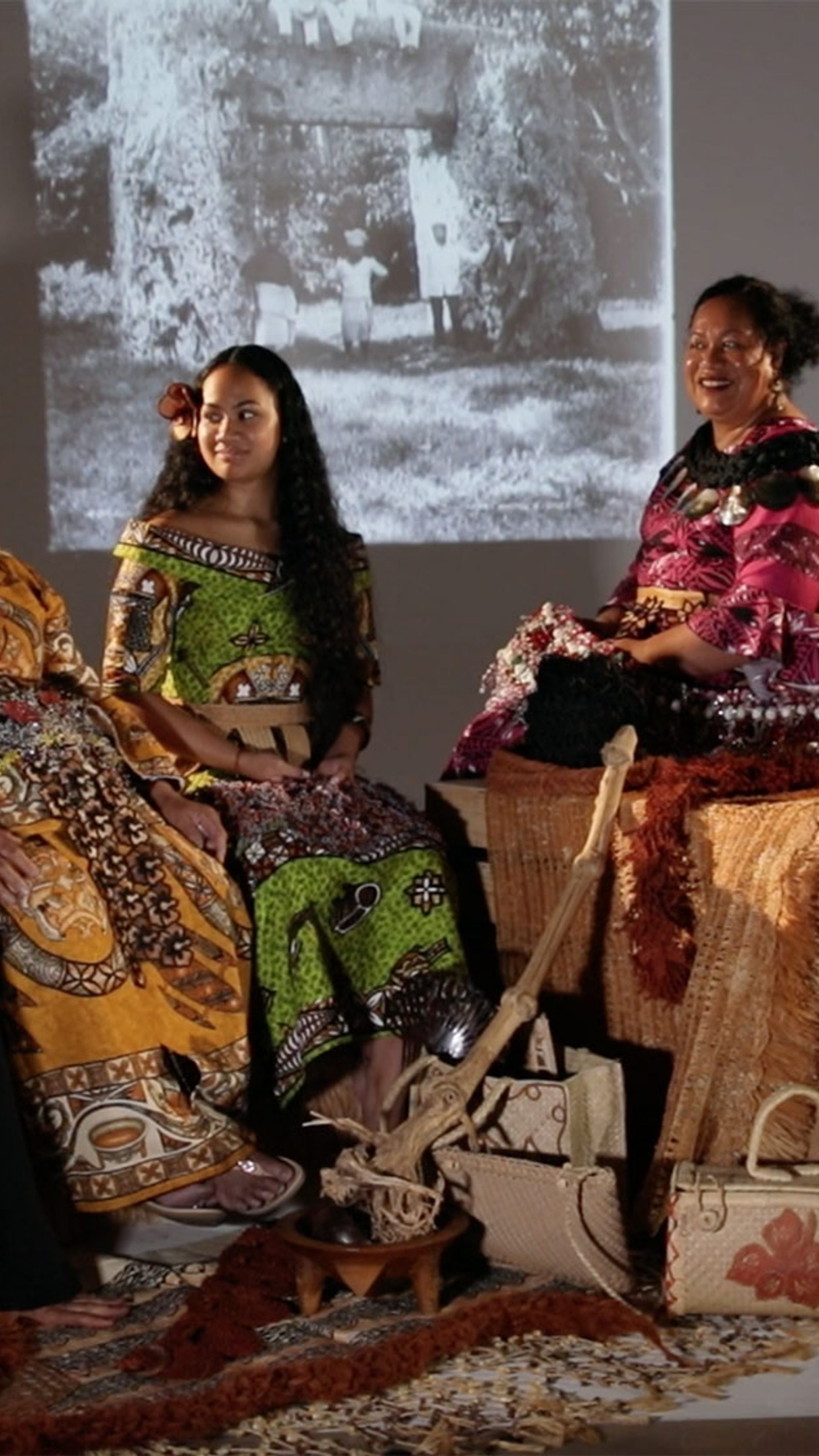 Four women and a man sitting in a semi-circle formation. One woman is sitting on the ground and the rest of the group are sitting on large boxes. They women are wearing traditional Tongan dress. The man is wearing traditional Samoan dress. They are holding a variety of objects.