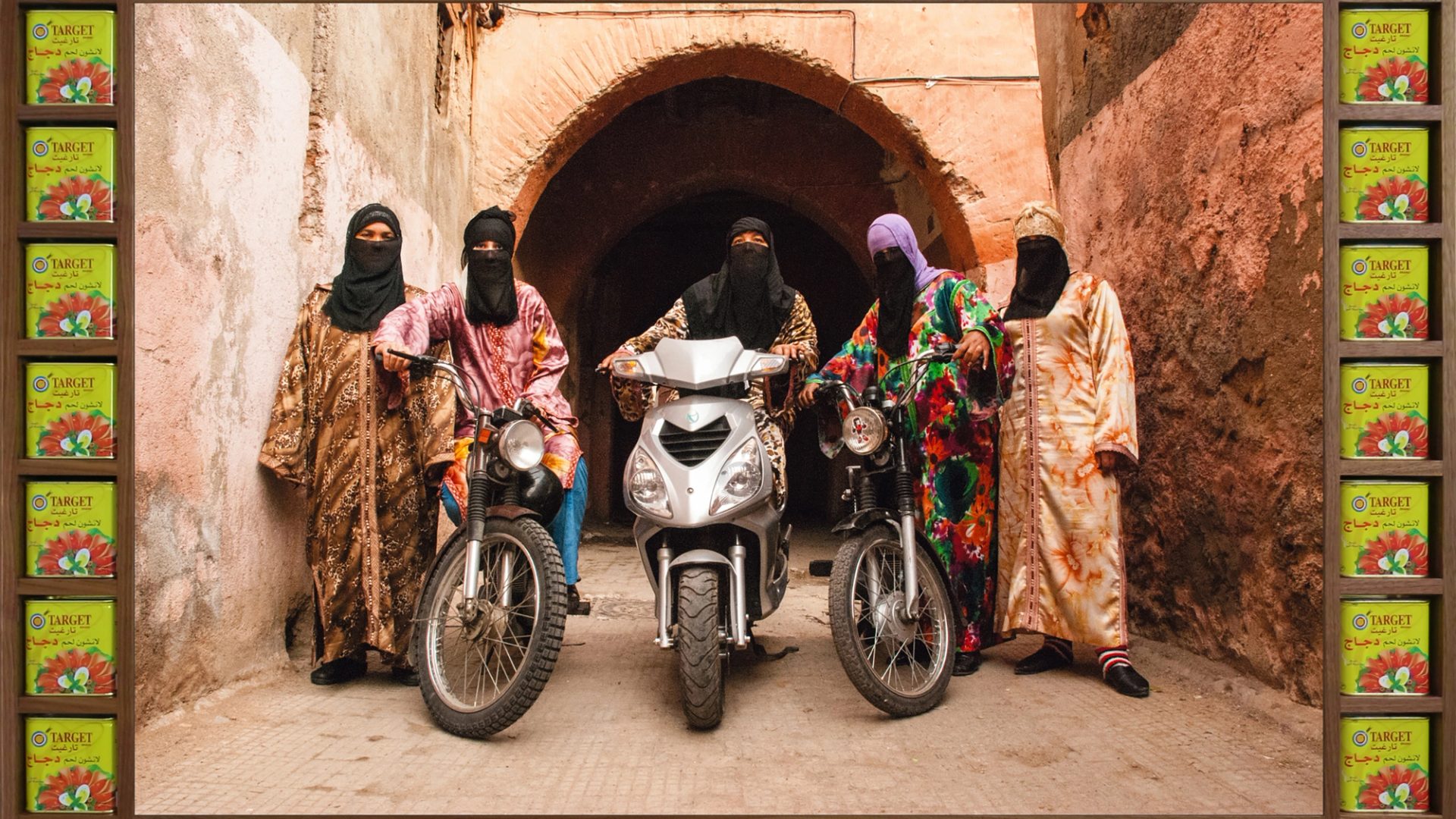 An art photograph that shows five figures in colourful Islamic-style dress with headscarves and black half niqabs. Three of the figures are astride motorcycles.