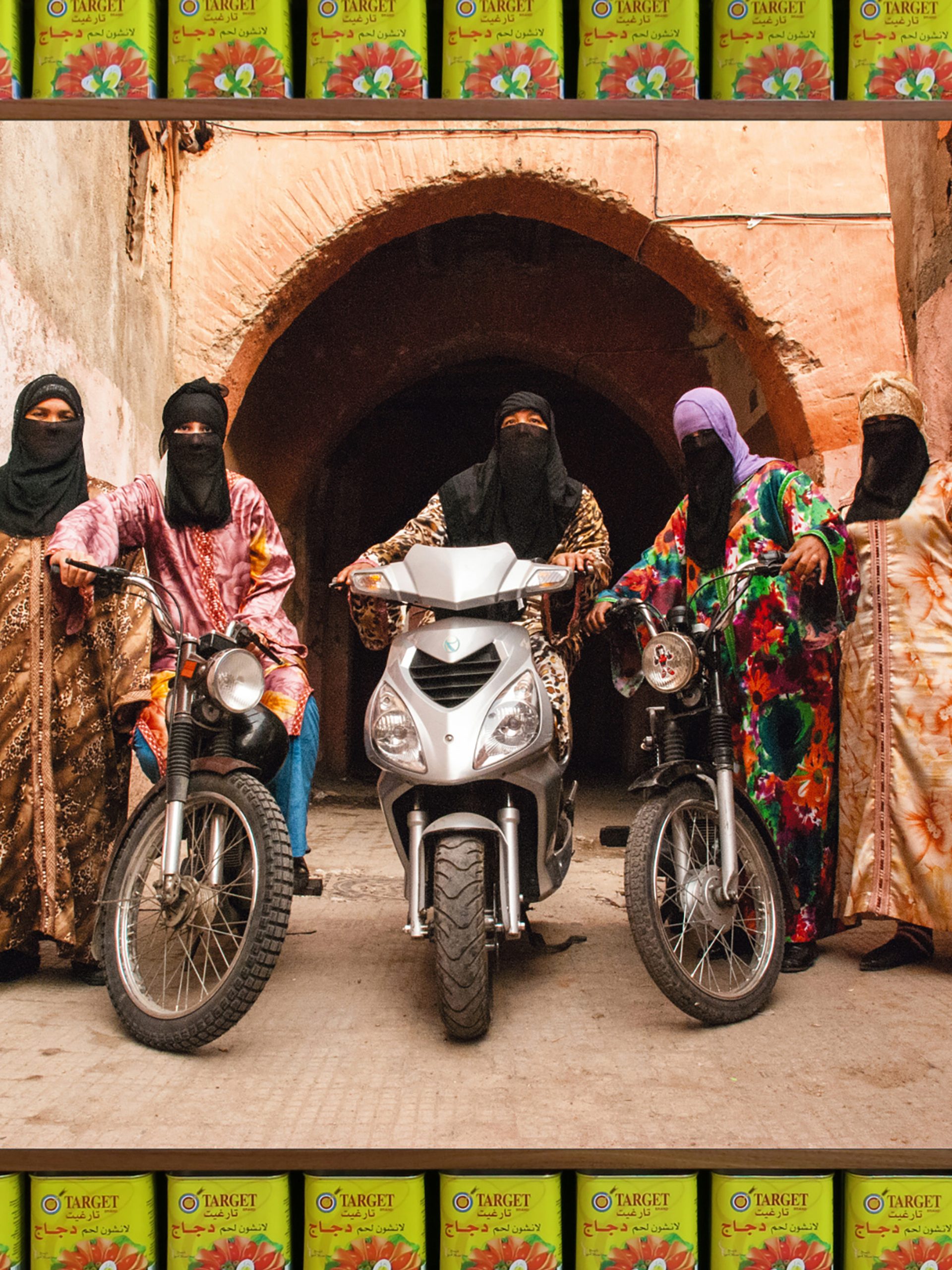 An art photograph that shows five figures in colourful Islamic-style dress with headscarves and black half niqabs. Three of the figures are astride motorcycles.