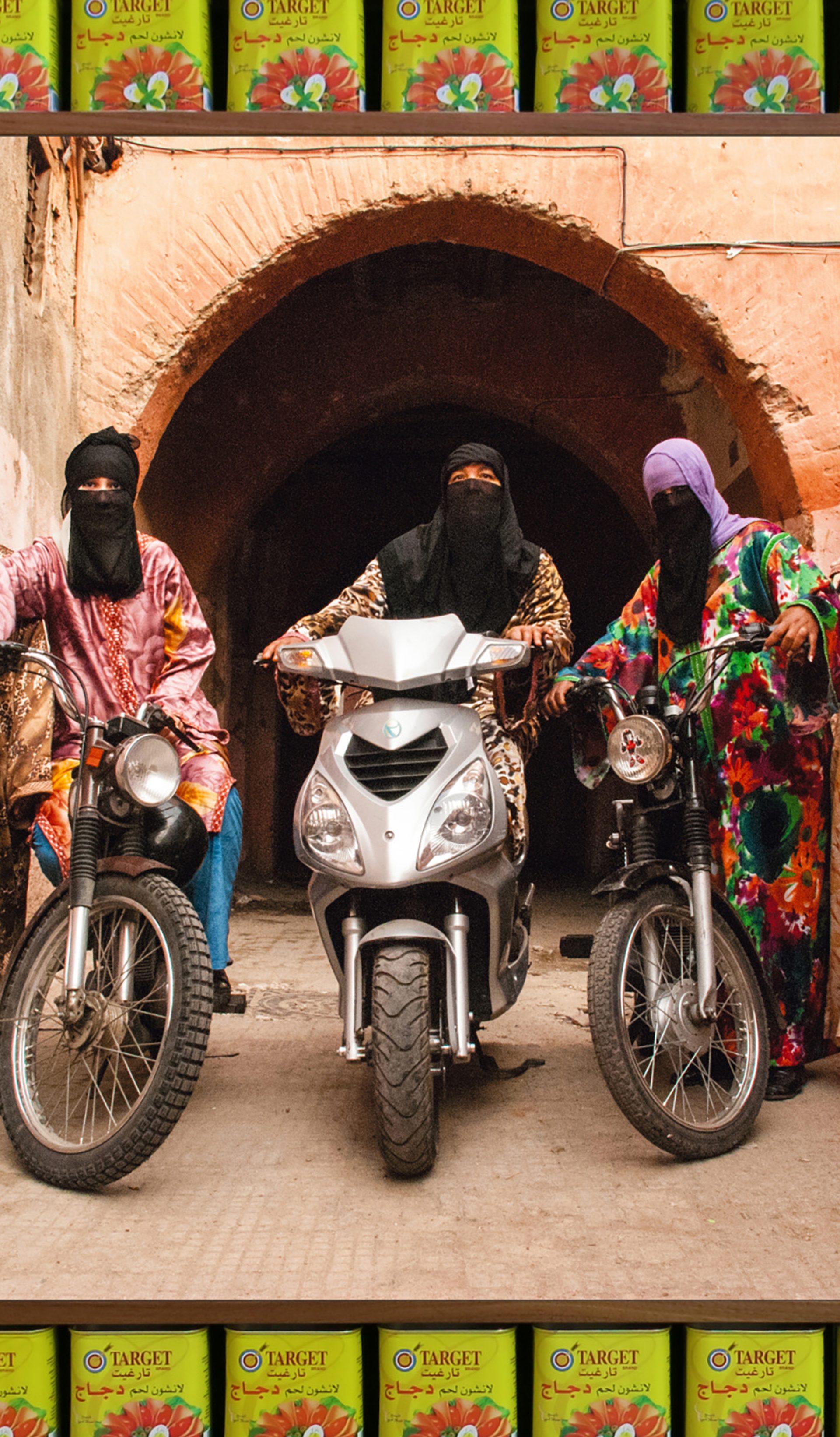 An art photograph that shows five figures in colourful Islamic-style dress with headscarves and black half niqabs. Three of the figures are astride motorcycles.