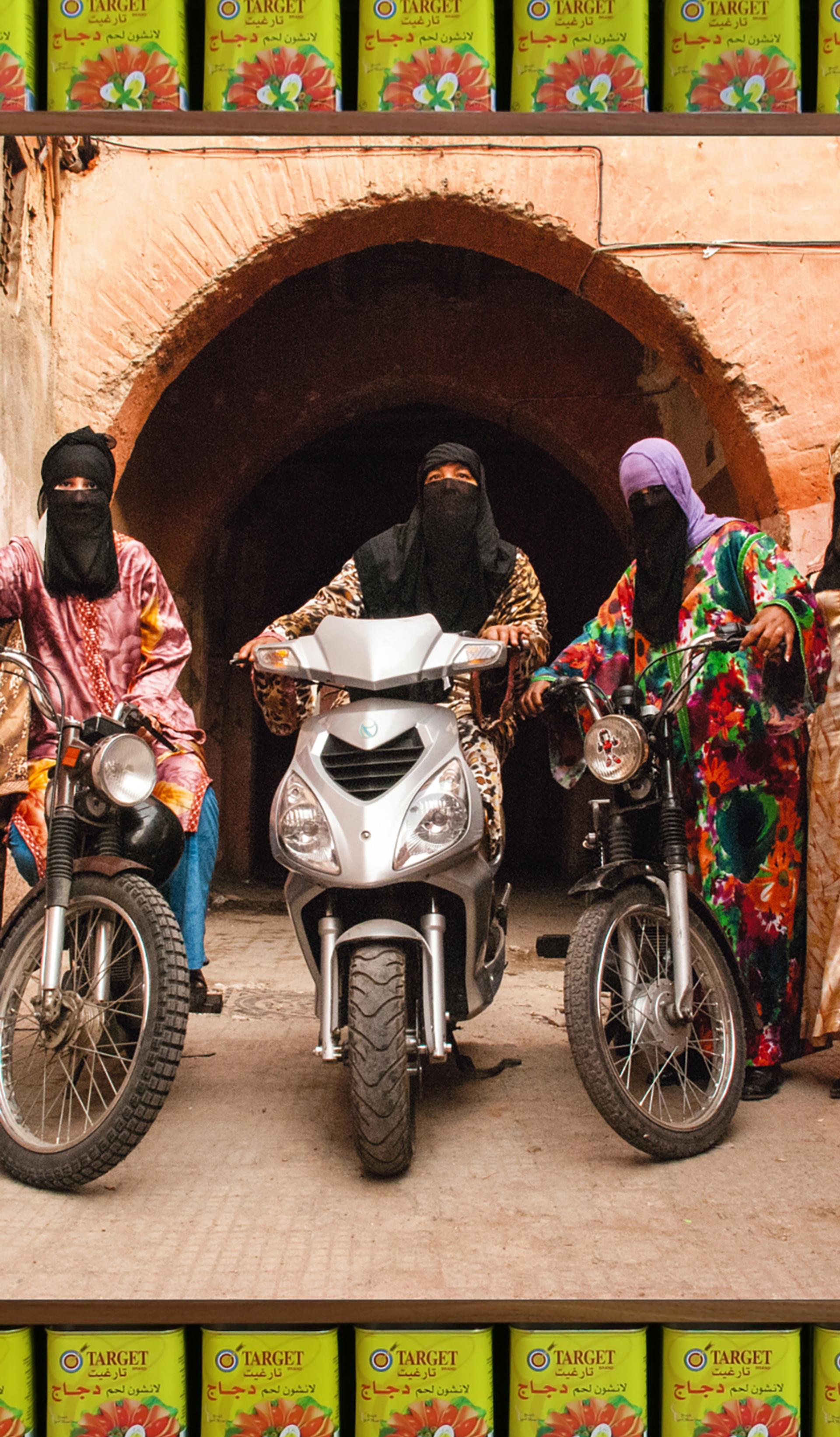 An art photograph that shows five figures in colourful Islamic-style dress with headscarves and black half niqabs. Three of the figures are astride motorcycles.
