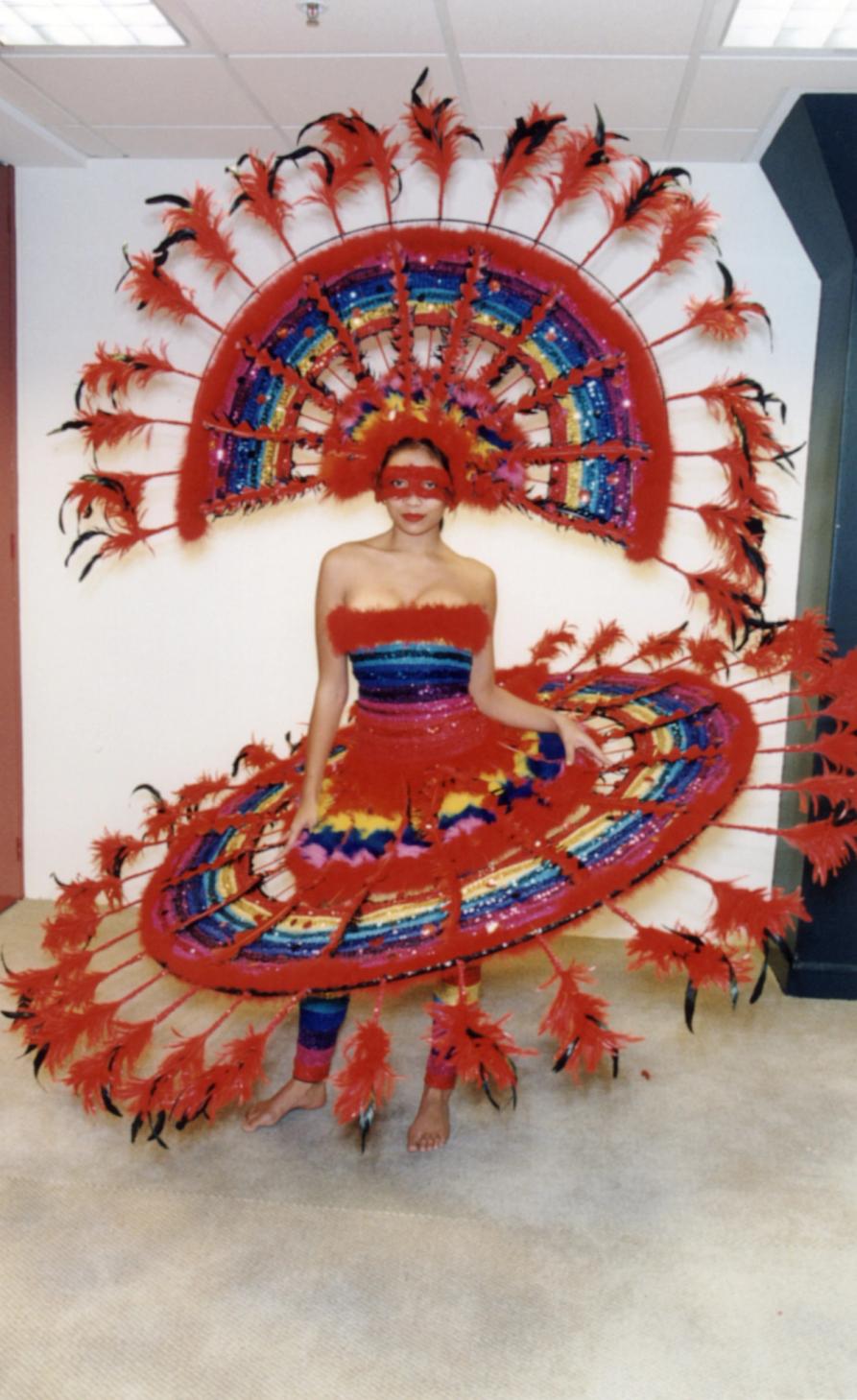 A woman dress in Rainbow dress with a stiff, circle shaped skirt and a large head dress with feathers.