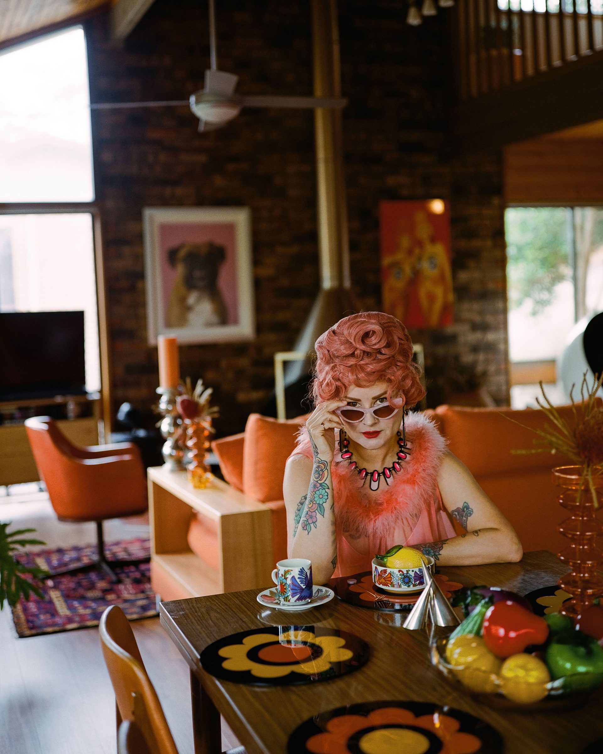 A woman with pink hair, wearing pink sunglasses and a pink fluffy top sits at a din