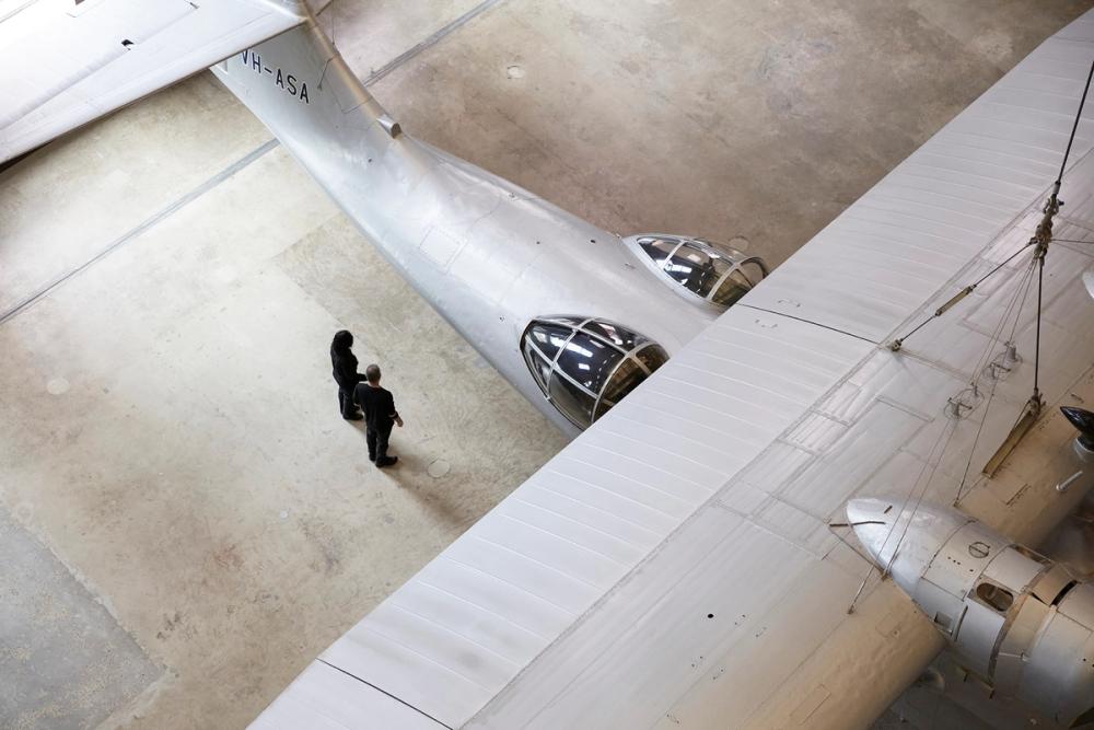 Partial aerial view of silver plane. Two people are standing close to the underneath of the wing.
