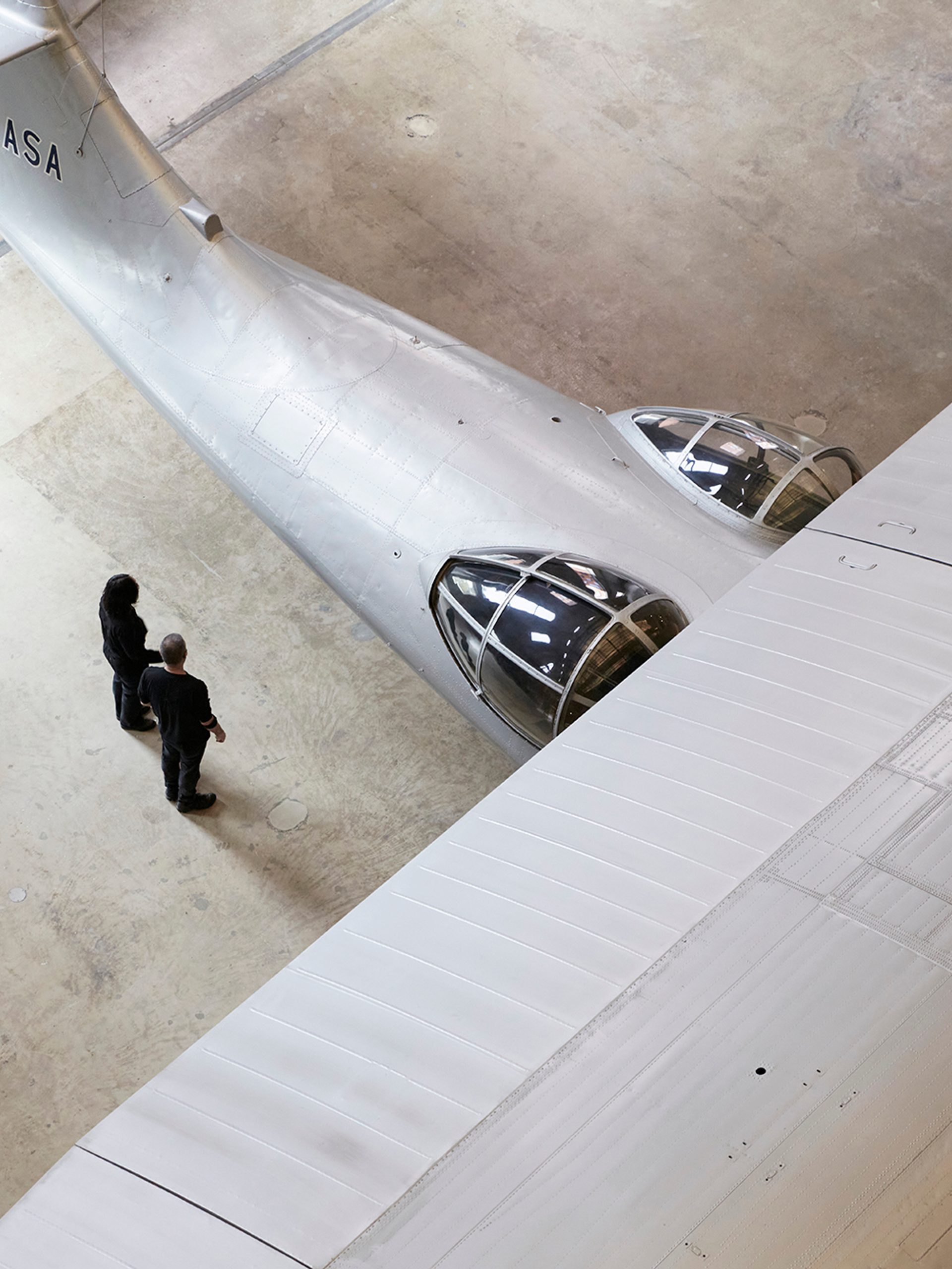 Partial aerial view of silver plane. Two people are standing close to the underneath of the wing.