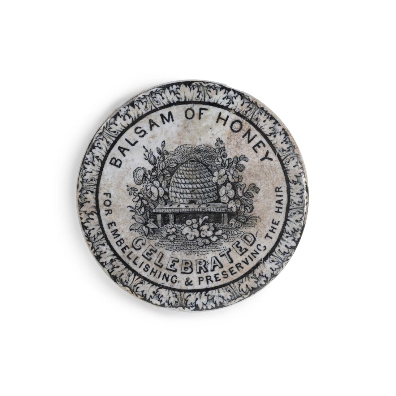 Ceramic lid of ‘Basalm of Honey’. Design includes floral border and in the centre, a beehive surrounded by flowers and plants.