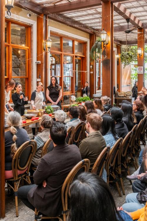 Crowd sits in an outdoor setting, watching chefs cook and talk to them.