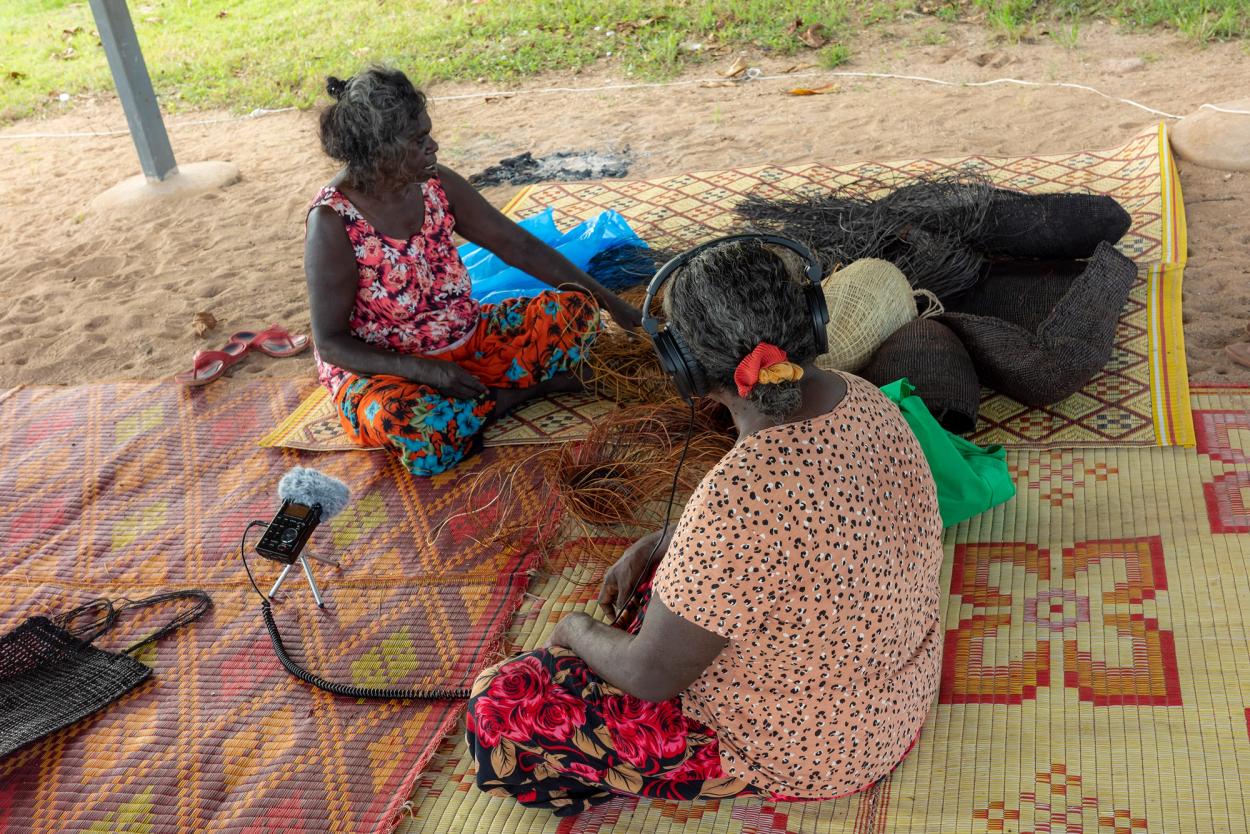Two women sitting outside on various woven mats.