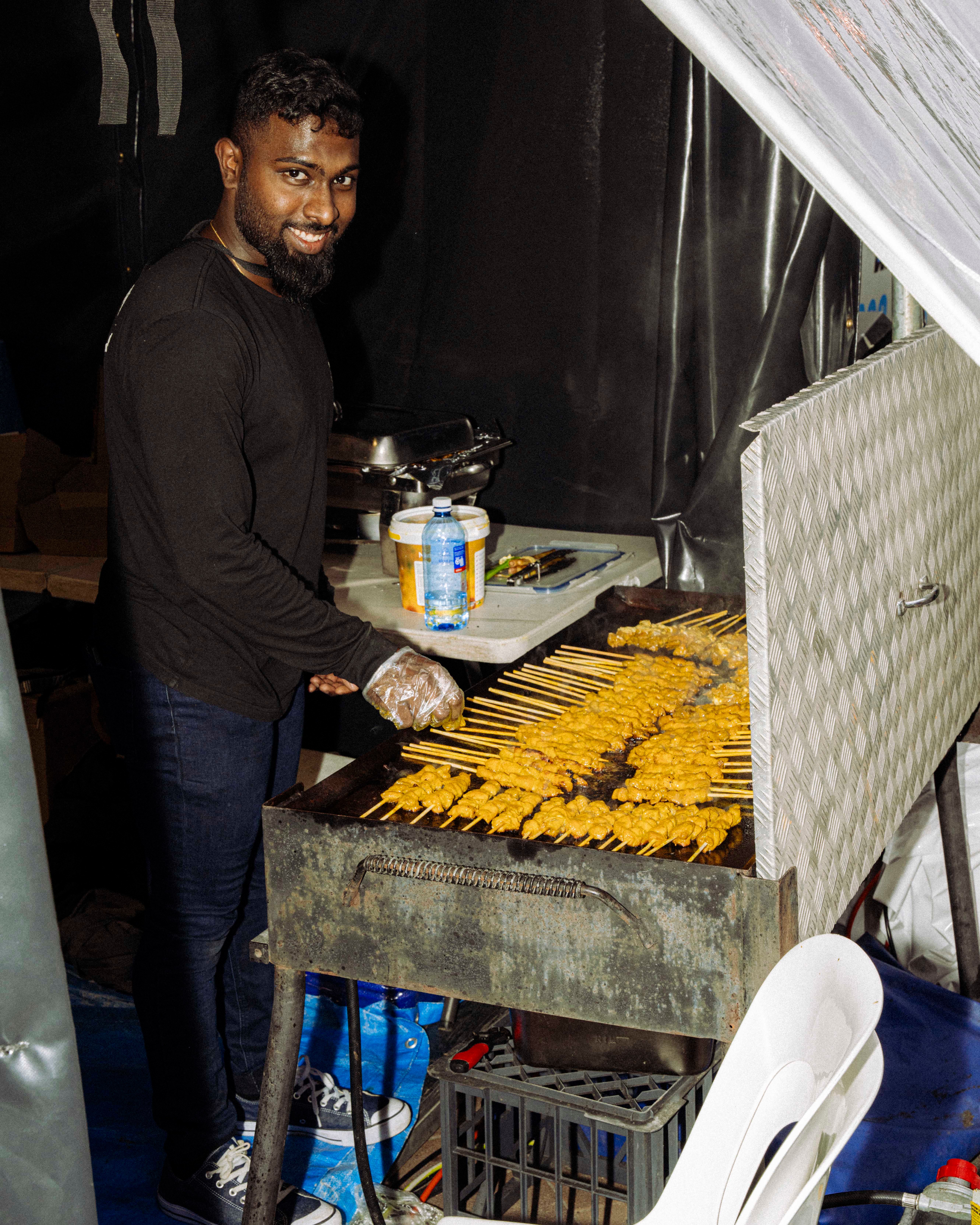 A smiling vendor in a black outfit with gloves stands at a grill, preparing food skewers of marinated meat, with sauce containers visible nearby.