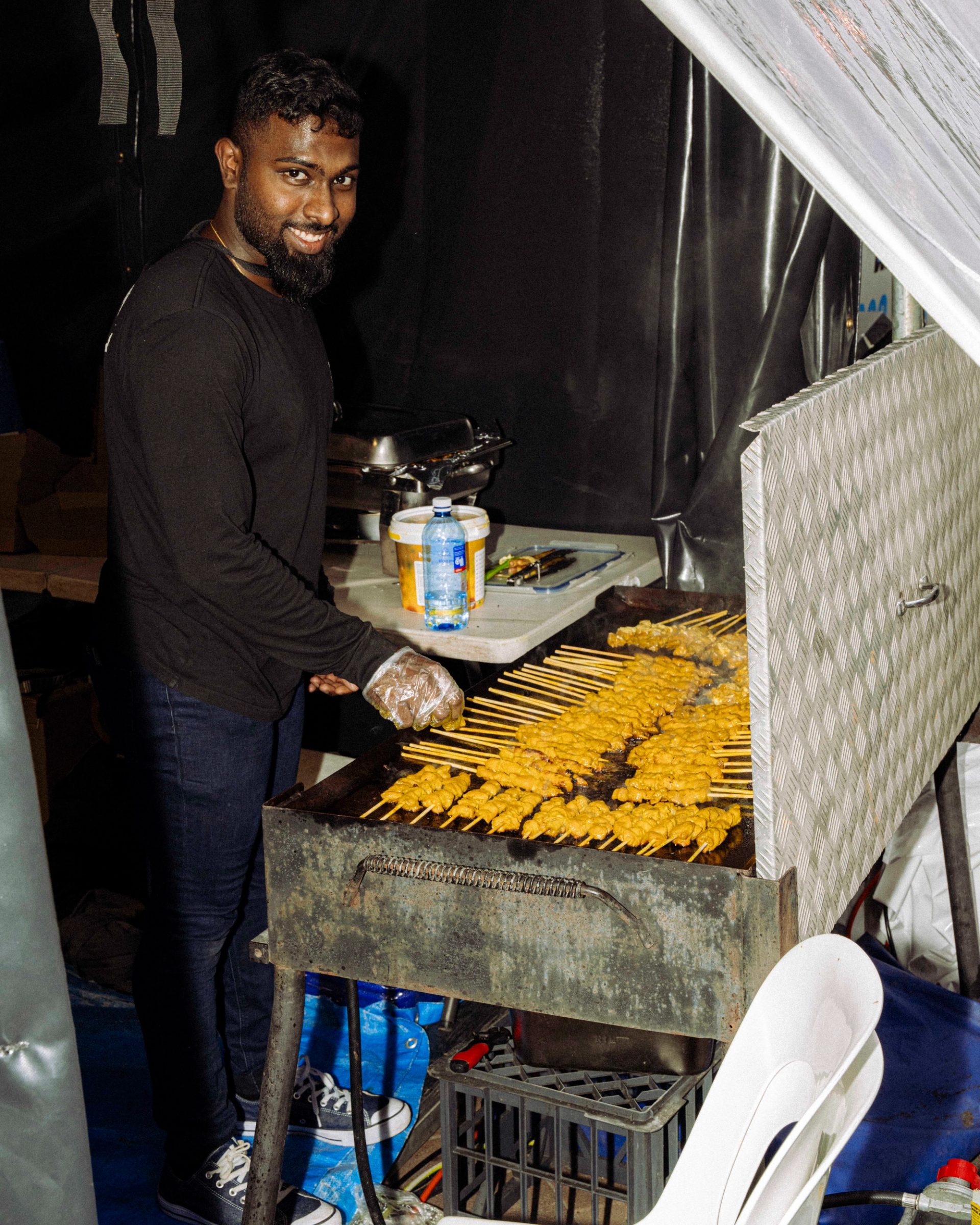 A smiling vendor in a black outfit with gloves stands at a grill, preparing food skewers of marinated meat, with sauce containers visible nearby.
