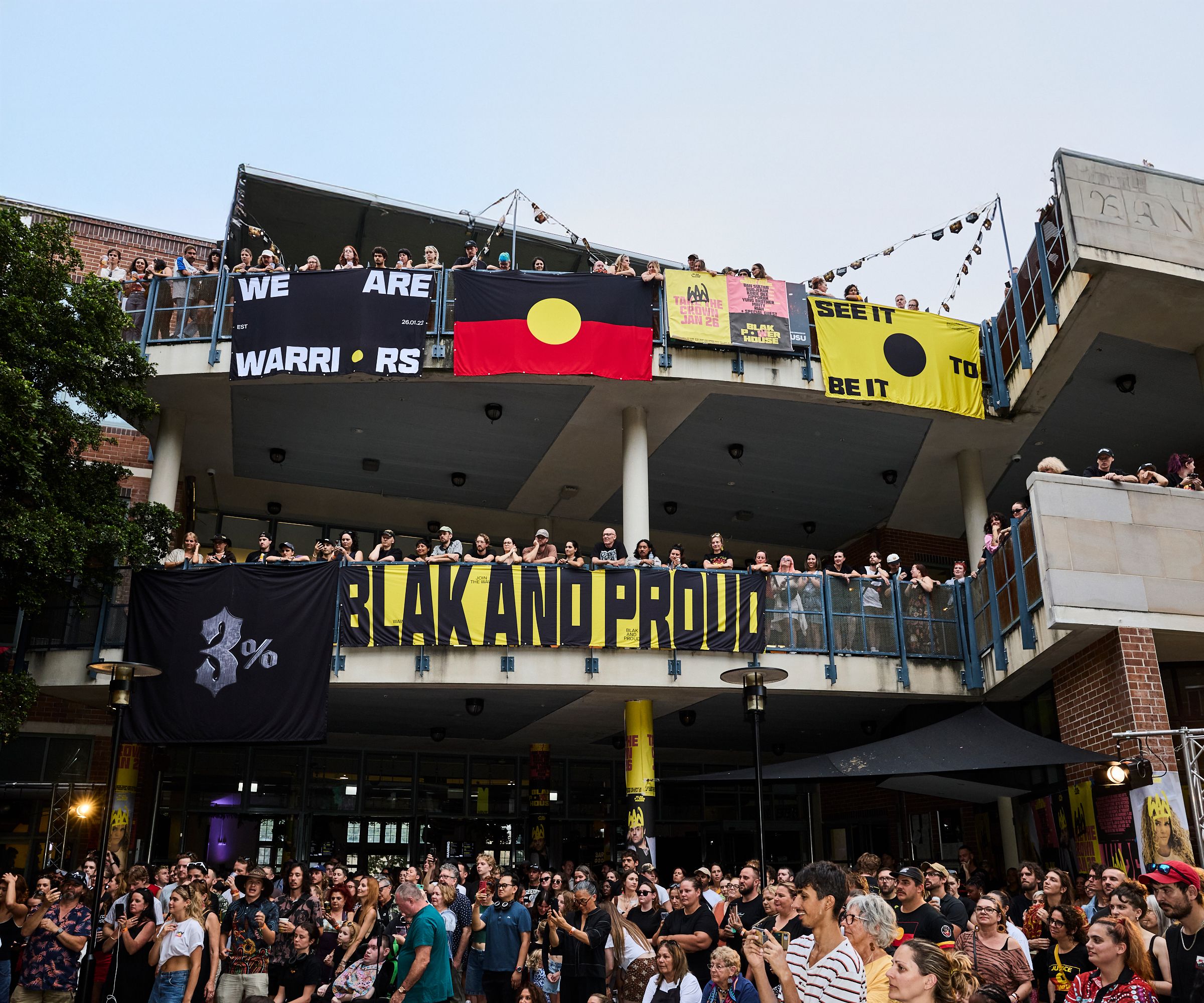 A photo looking up to a two level building, crowds filling the balconies. Bright posters and the Aboriginal flag hangs over the edges, while the crowd looks to the left watching a stage show.