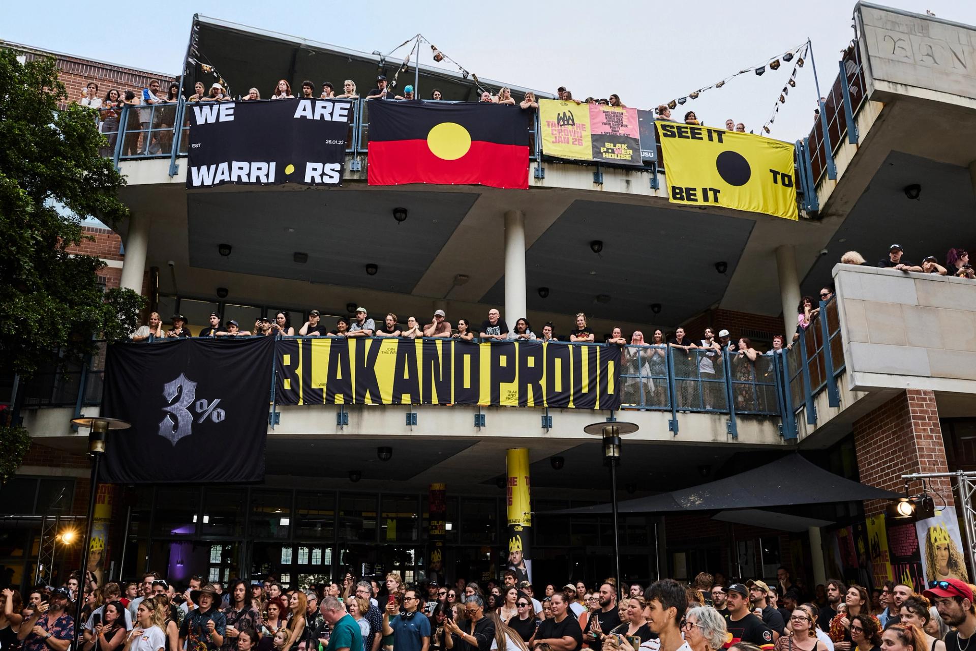 A photo looking up to a two level building, crowds filling the balconies. Bright posters and the Aboriginal flag hangs over the edges, while the crowd looks to the left watching a stage show.