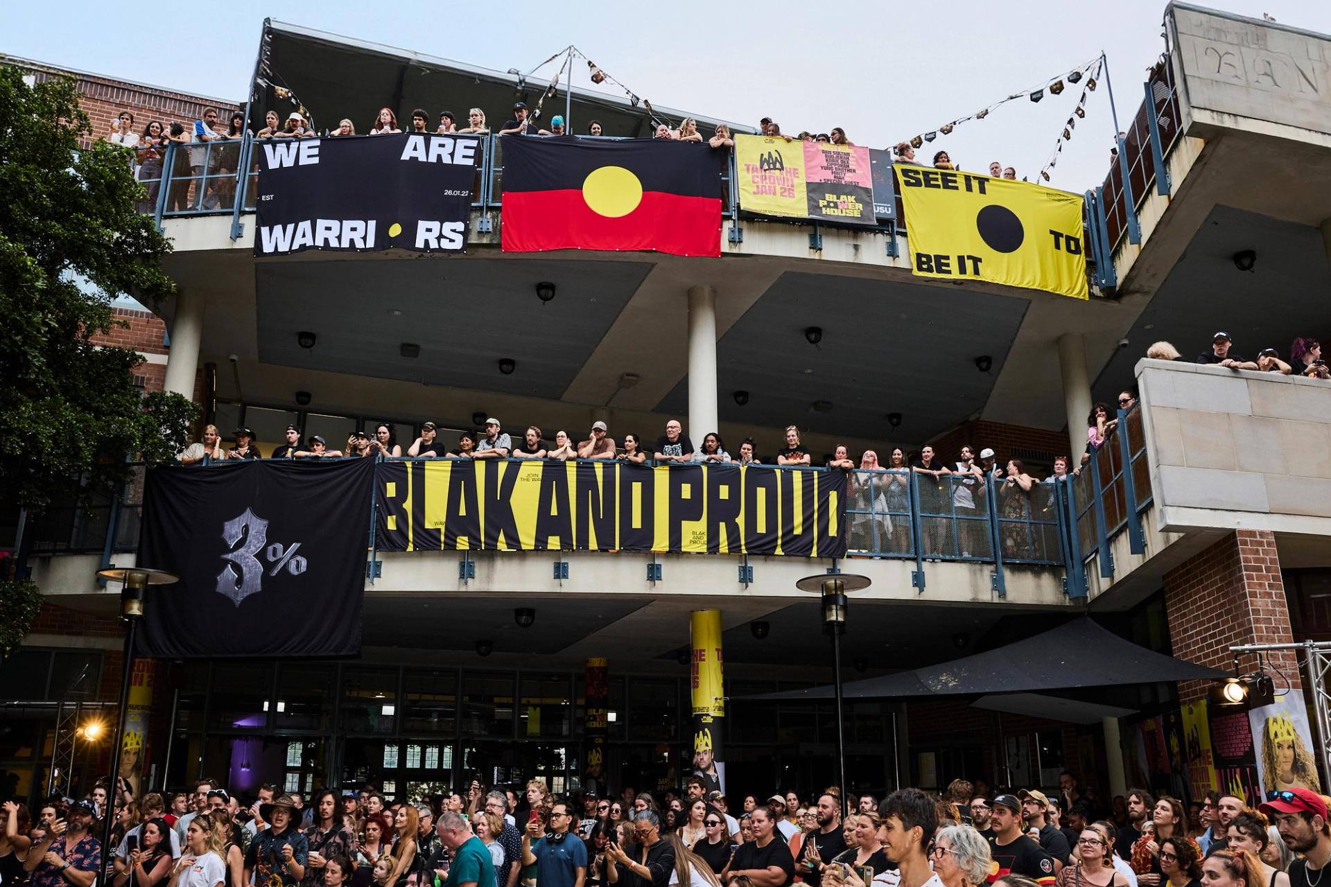 A photo looking up to a two level building, crowds filling the balconies. Bright posters and the Aboriginal flag hangs over the edges, while the crowd looks to the left watching a stage show.