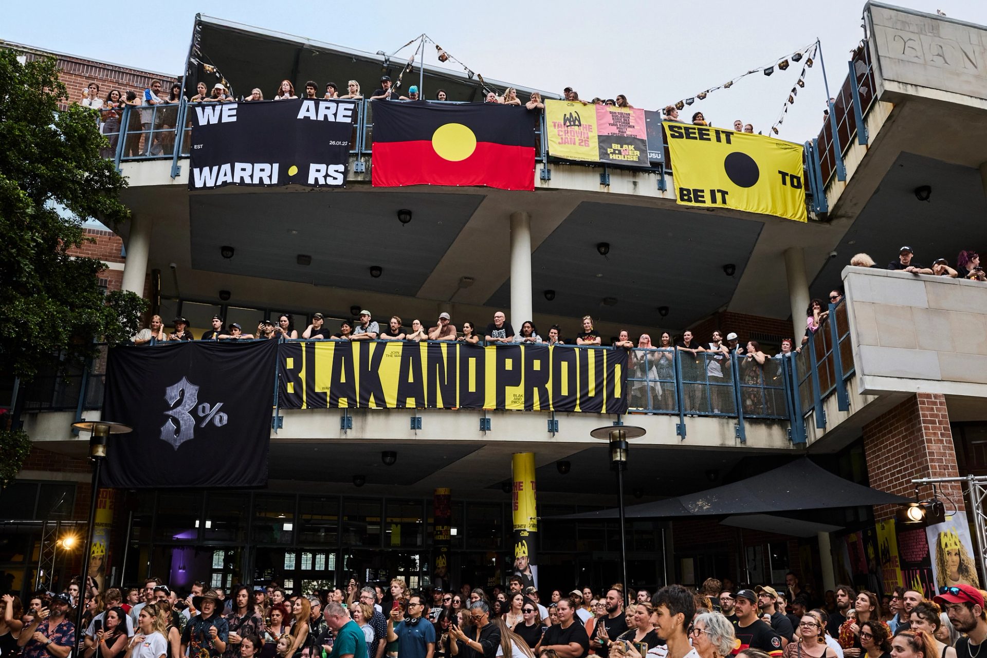 A photo looking up to a two level building, crowds filling the balconies. Bright posters and the Aboriginal flag hangs over the edges, while the crowd looks to the left watching a stage show.