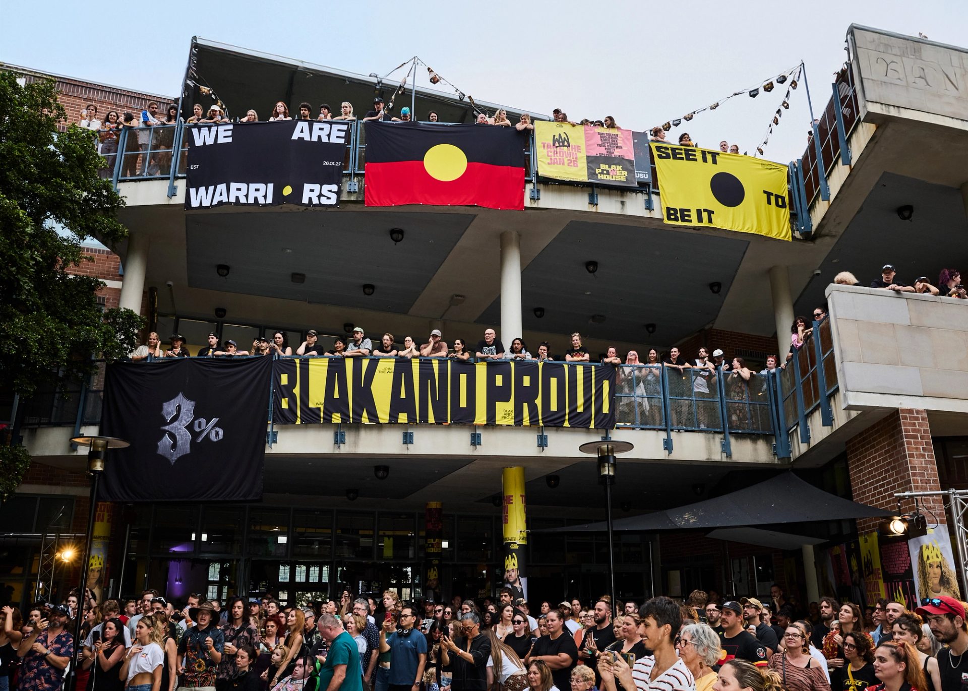 A photo looking up to a two level building, crowds filling the balconies. Bright posters and the Aboriginal flag hangs over the edges, while the crowd looks to the left watching a stage show.